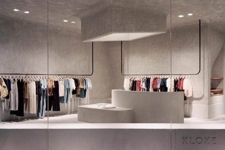 Modern clothing boutique with racks of assorted clothes, minimalist concrete interior, and a curved counter