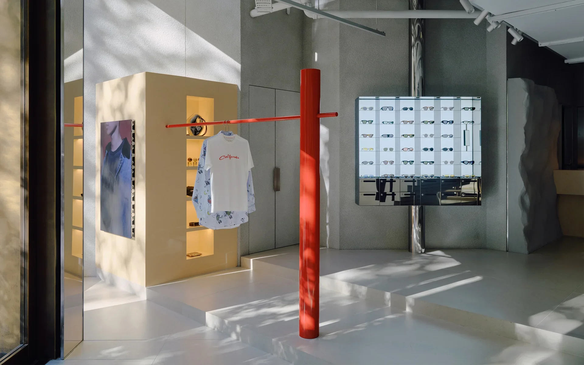 Interior of a modern eyewear store with sunglasses display, clothing item on a red pole, and abstract wall decor.
