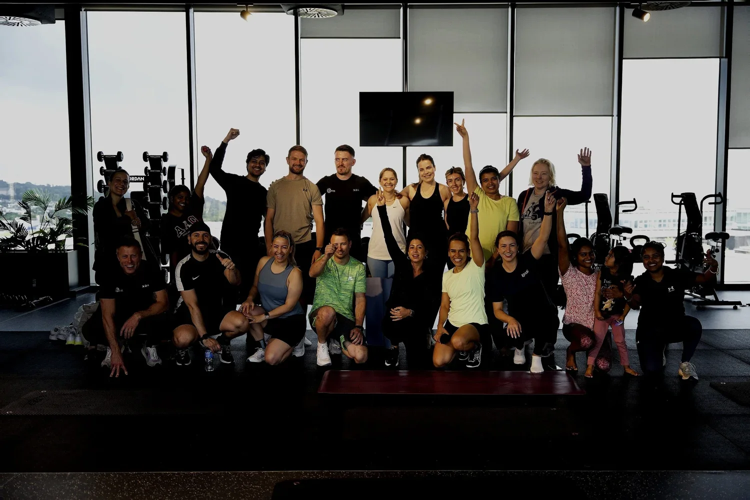 A large group of people posing together at a gym, some sitting and some standing, with gym equipment and large windows in the background.
