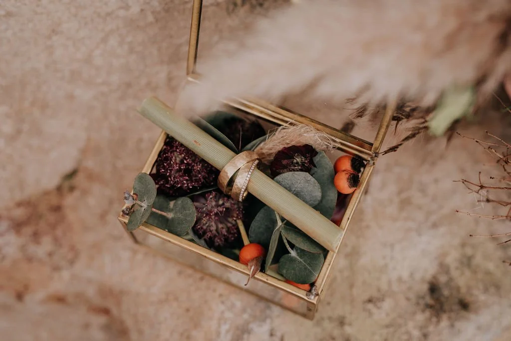 Open wooden box with dried plants, stones, and orange berries, with a stick and a ribbon inside.