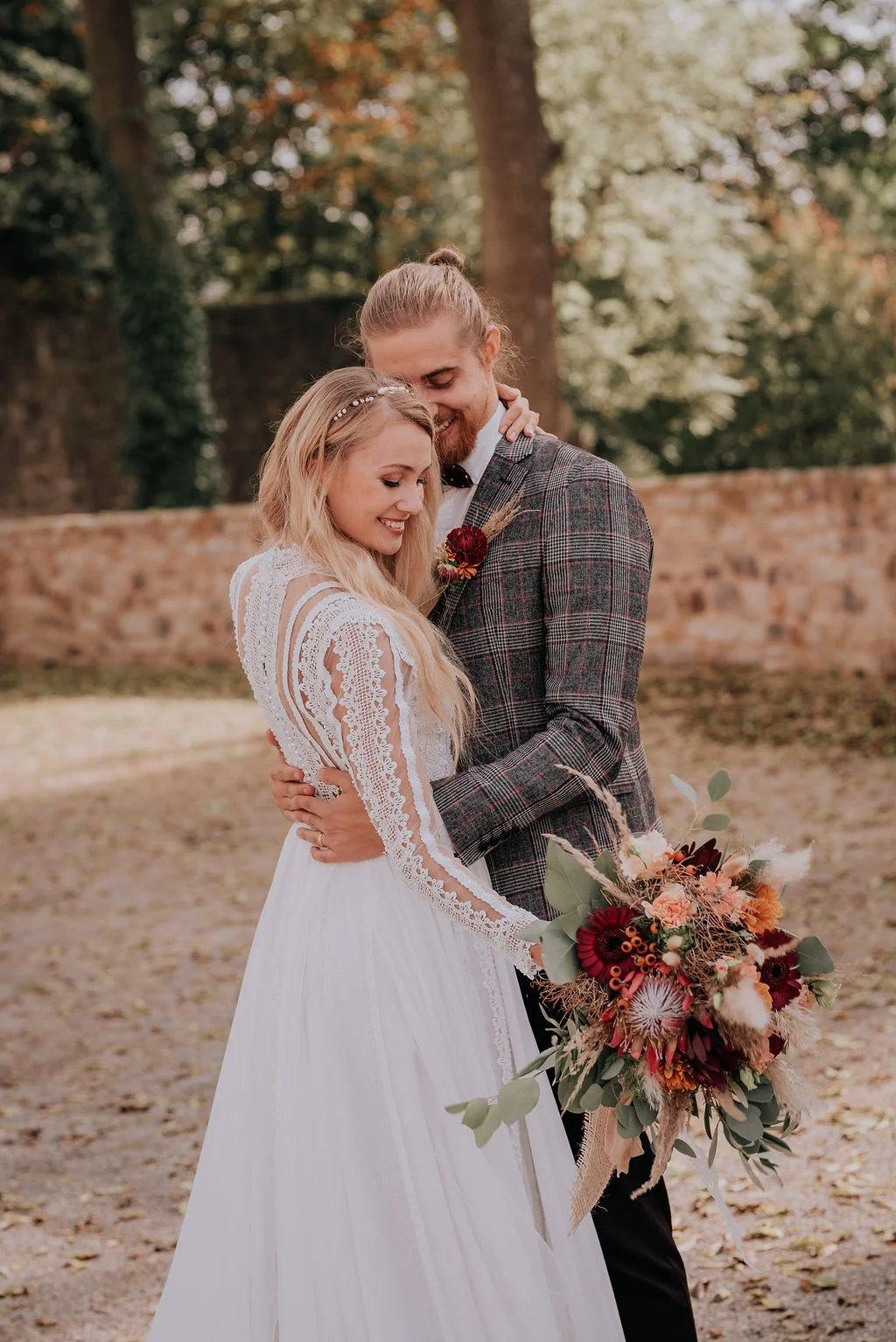 A bride and groom embrace outdoors, smiling and looking down, with the bride holding a large bouquet of flowers, during daytime.