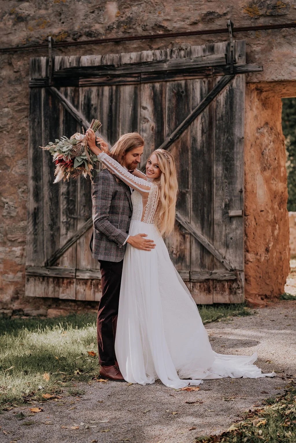 A bride and groom embrace outdoors in front of a rustic wooden barn door, the bride holding a bouquet of flowers above the groom's head and smiling at the camera.