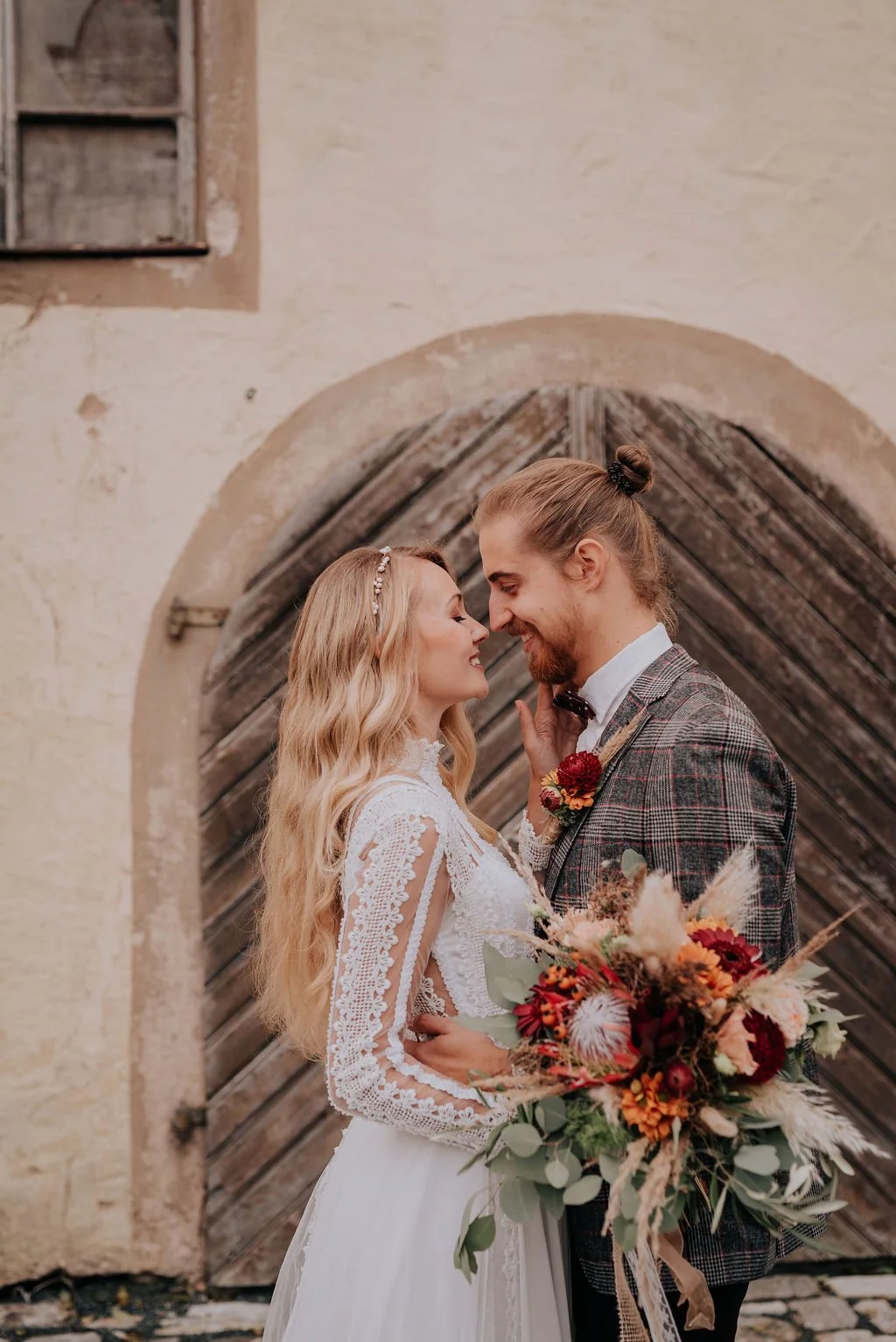 A bride and groom face each other in front of a rustic wooden door, holding a bouquet with red, orange, and beige flowers. The bride wears a white lace wedding dress, and the groom is dressed in a plaid suit with a boutonniere.