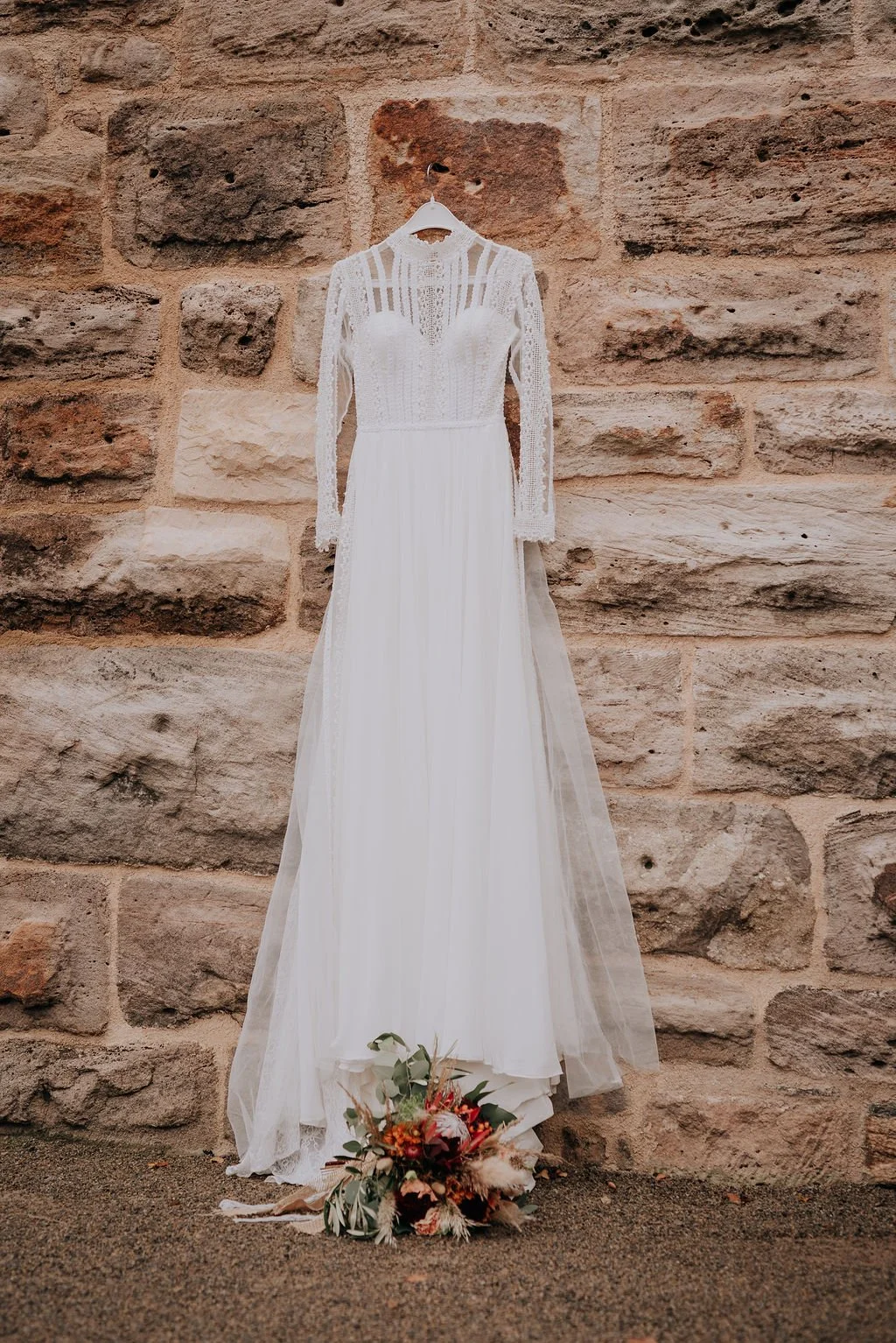 A white wedding dress hanging on a hanger against a stone wall, with a bouquet of flowers on the ground in front of it.