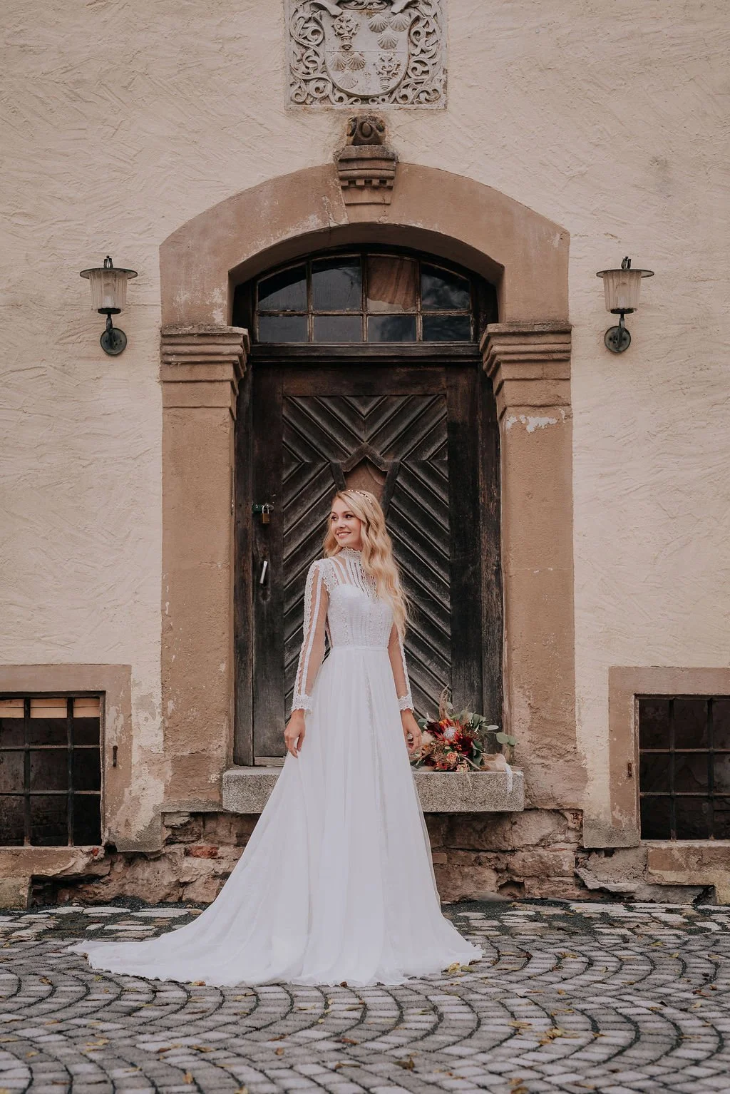 A woman in a white wedding dress standing outside in front of an old wooden door with stone framing, holding a bouquet, with a smile on her face.