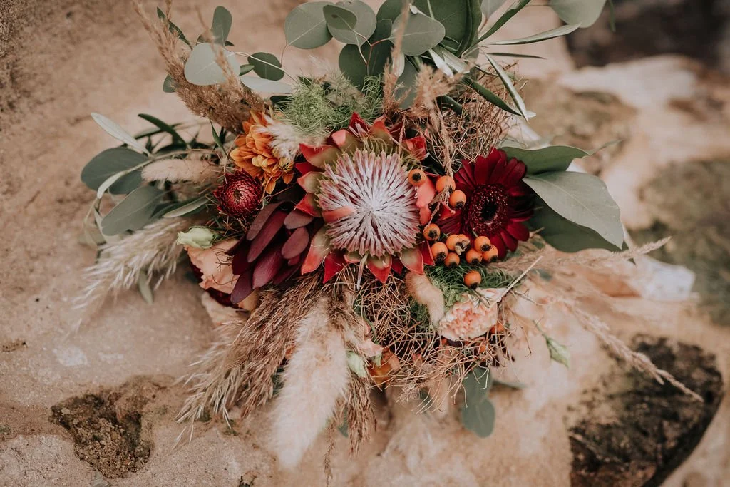 A bouquet of dried flowers and foliage with various textures and colors, resting on sandy ground.