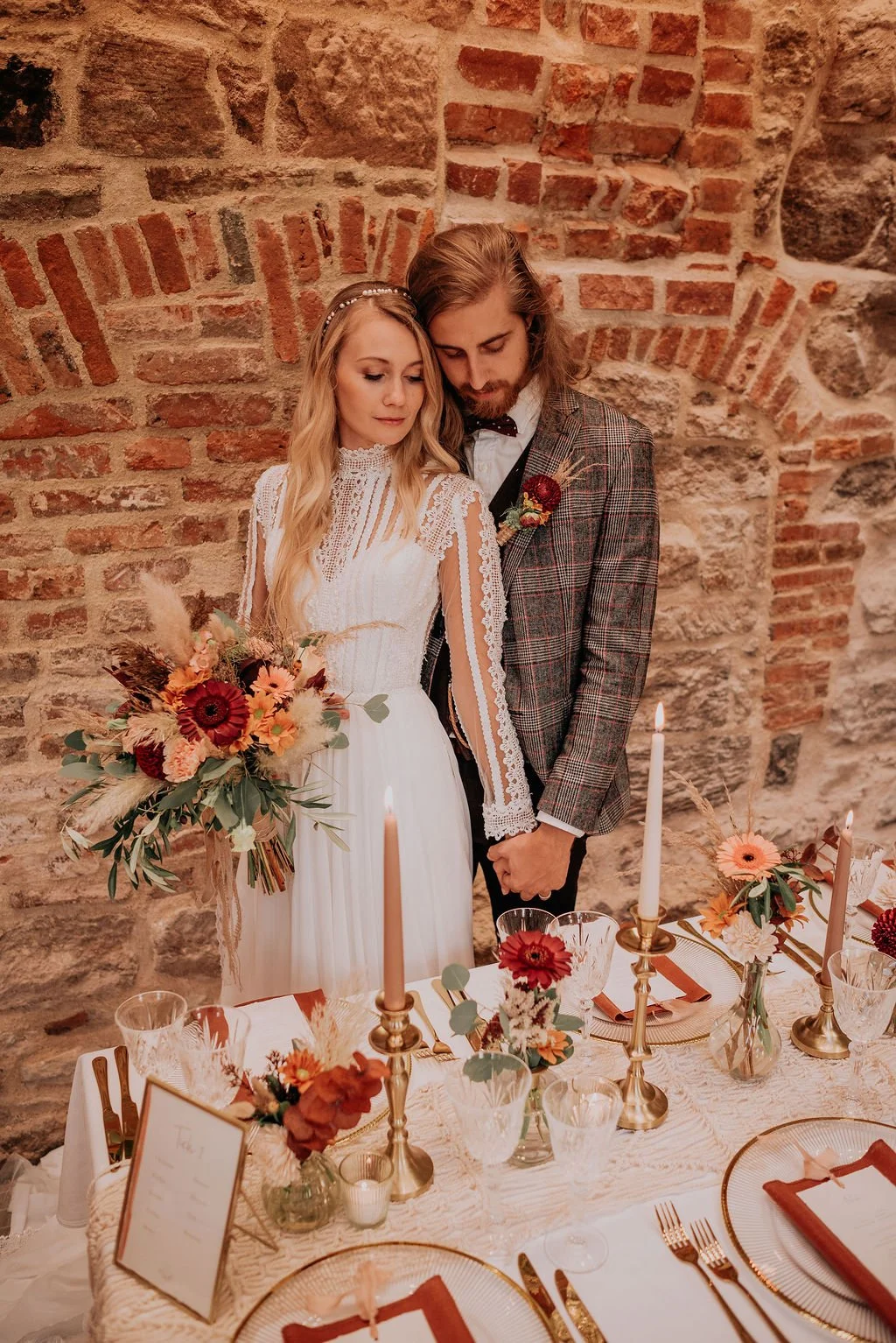 A bride and groom standing close together at a wedding reception table, holding hands and looking down. The bride is dressed in a white lace wedding gown, holding a bouquet of flowers with orange, red, and green tones. The groom is wearing a plaid su