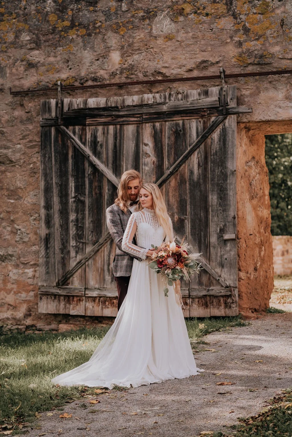 A bride and groom standing close together outdoors beside a rustic wooden barn door, with the bride holding a bouquet of flowers, both dressed in wedding attire.