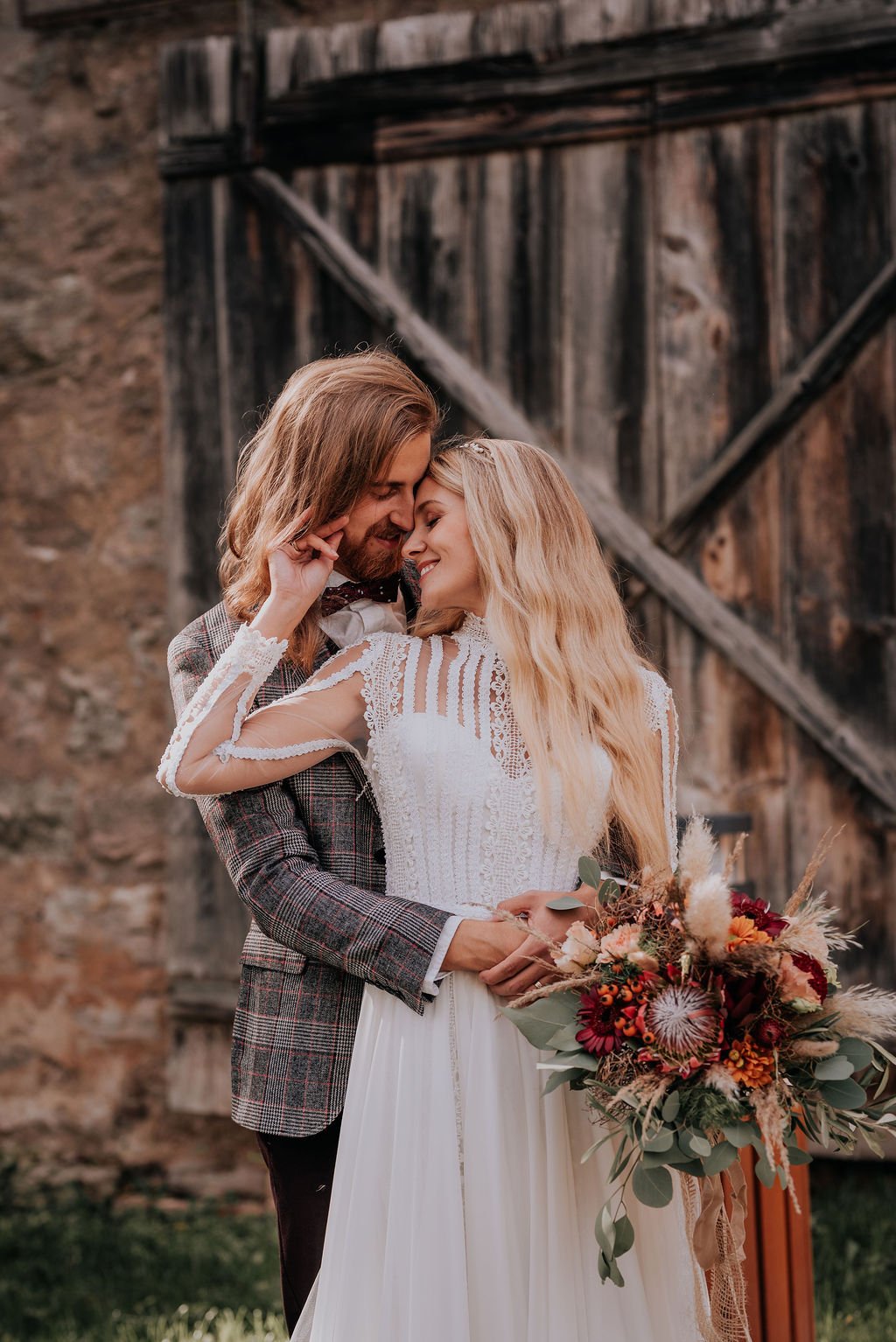A bride and groom share a romantic moment outdoors during their wedding, with the bride holding a bouquet of flowers and dressed in a detailed white wedding dress, and the groom in a plaid jacket.