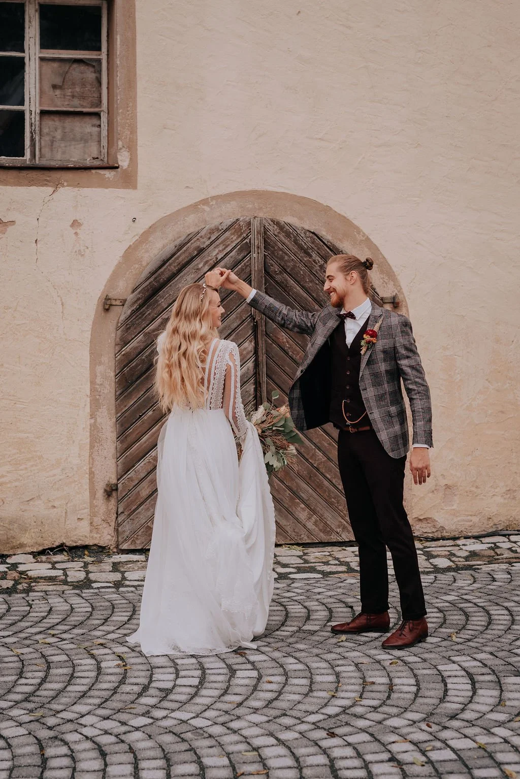 A couple dressed in wedding attire dancing in front of a rustic wooden door, with the groom twirling the bride.