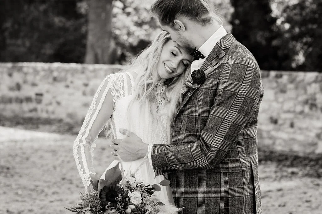 A black and white photograph of a happy bride and groom on their wedding day, embracing outdoors, with the bride holding a bouquet of flowers.