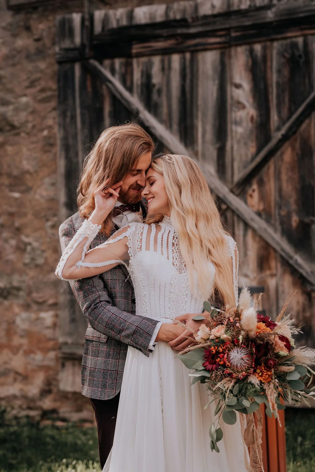 A couple dressed in wedding attire sharing a romantic moment outdoors, with a rustic wooden barn in the background. The woman is holding a bouquet of flowers.