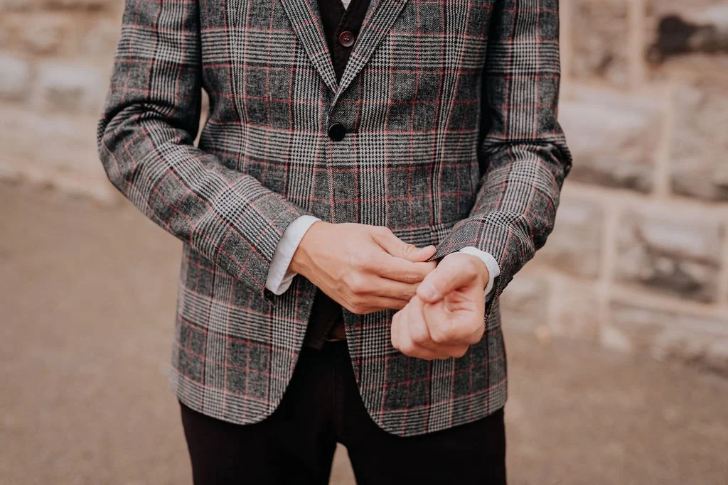 Person adjusting the sleeve of a plaid blazer against a brick wall background.