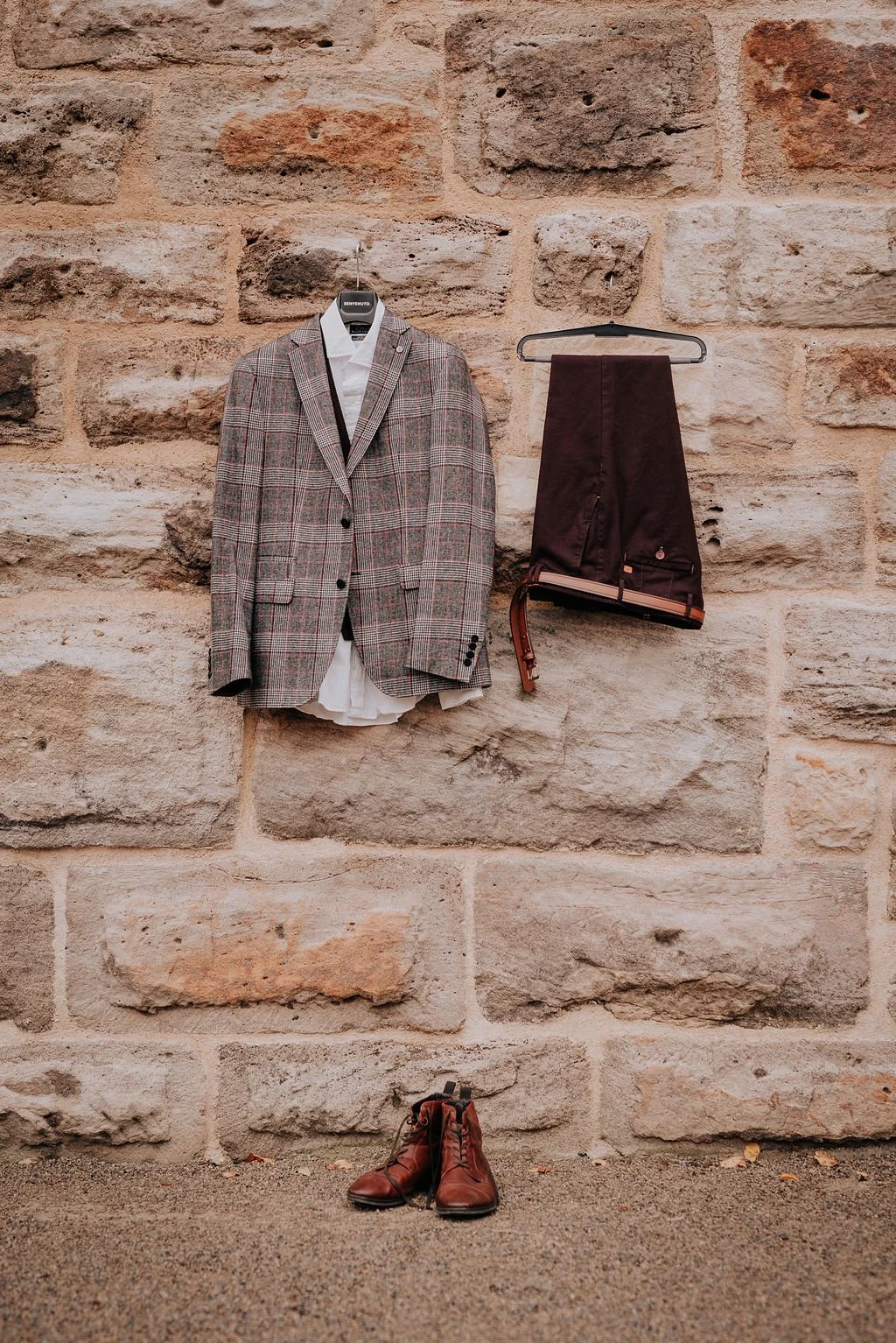 A brick wall with a plaid blazer, white shirt, dark pants, a belt, and brown dress shoes arranged against it.