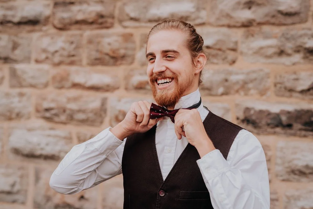 A man with a beard and long hair tied back, smiling, adjusting his bow tie, wearing a white shirt and black vest, in front of a brick wall.