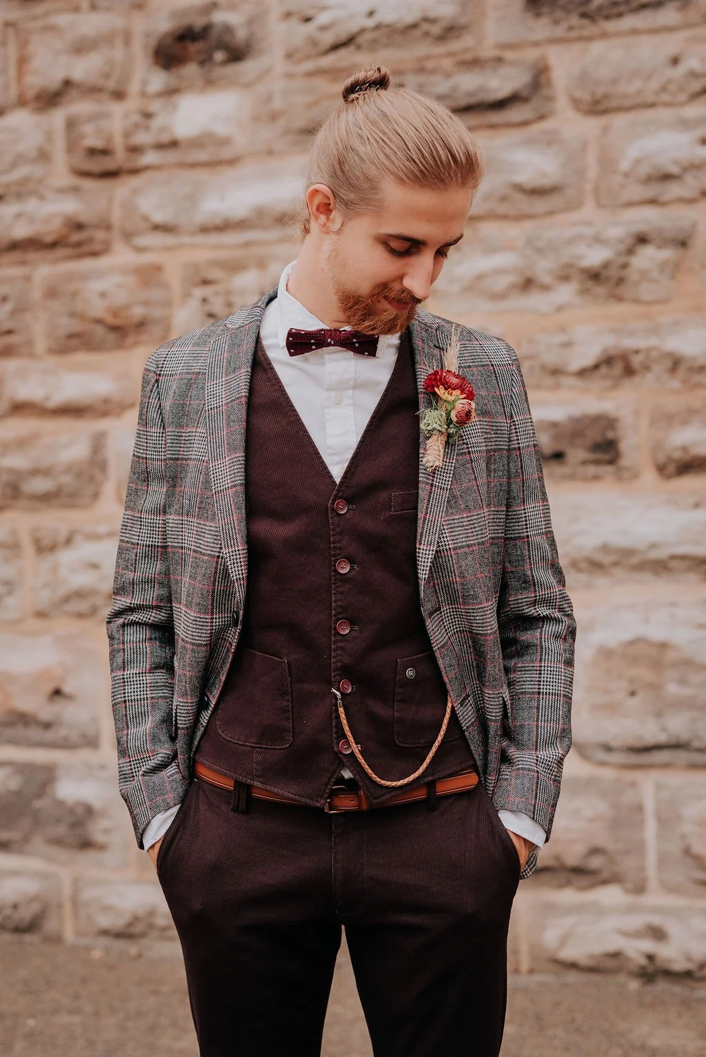 A man in formal attire with a plaid blazer, dark vest, and bow tie, standing against a stone wall, with a boutonniere pinned to his blazer.