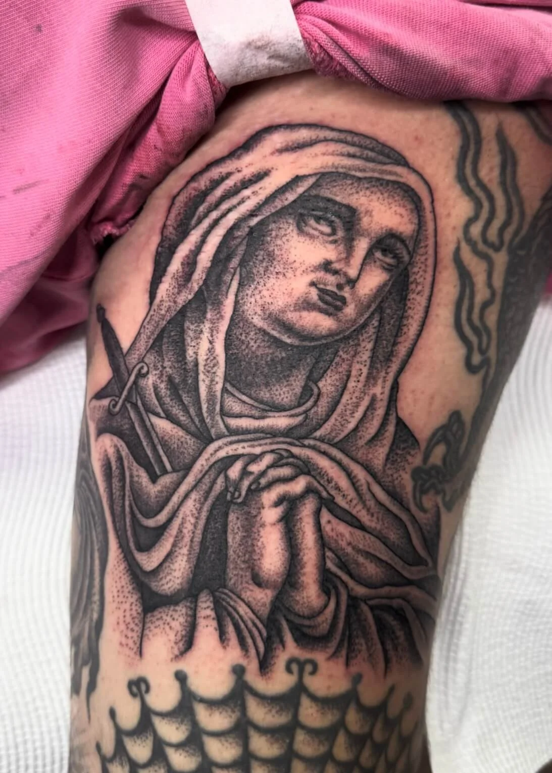 Tattoo of a praying woman with a halo and long hair, holding her hands together. The tattoo is in black and gray ink with detailed shading.