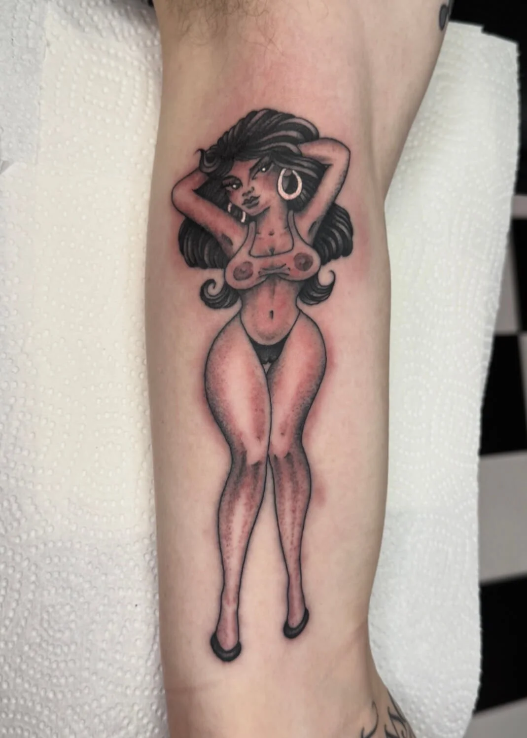 Tattoo of a woman with long flowing hair, large hoop earrings, and a necklace, standing with legs together and arms raised behind her head.