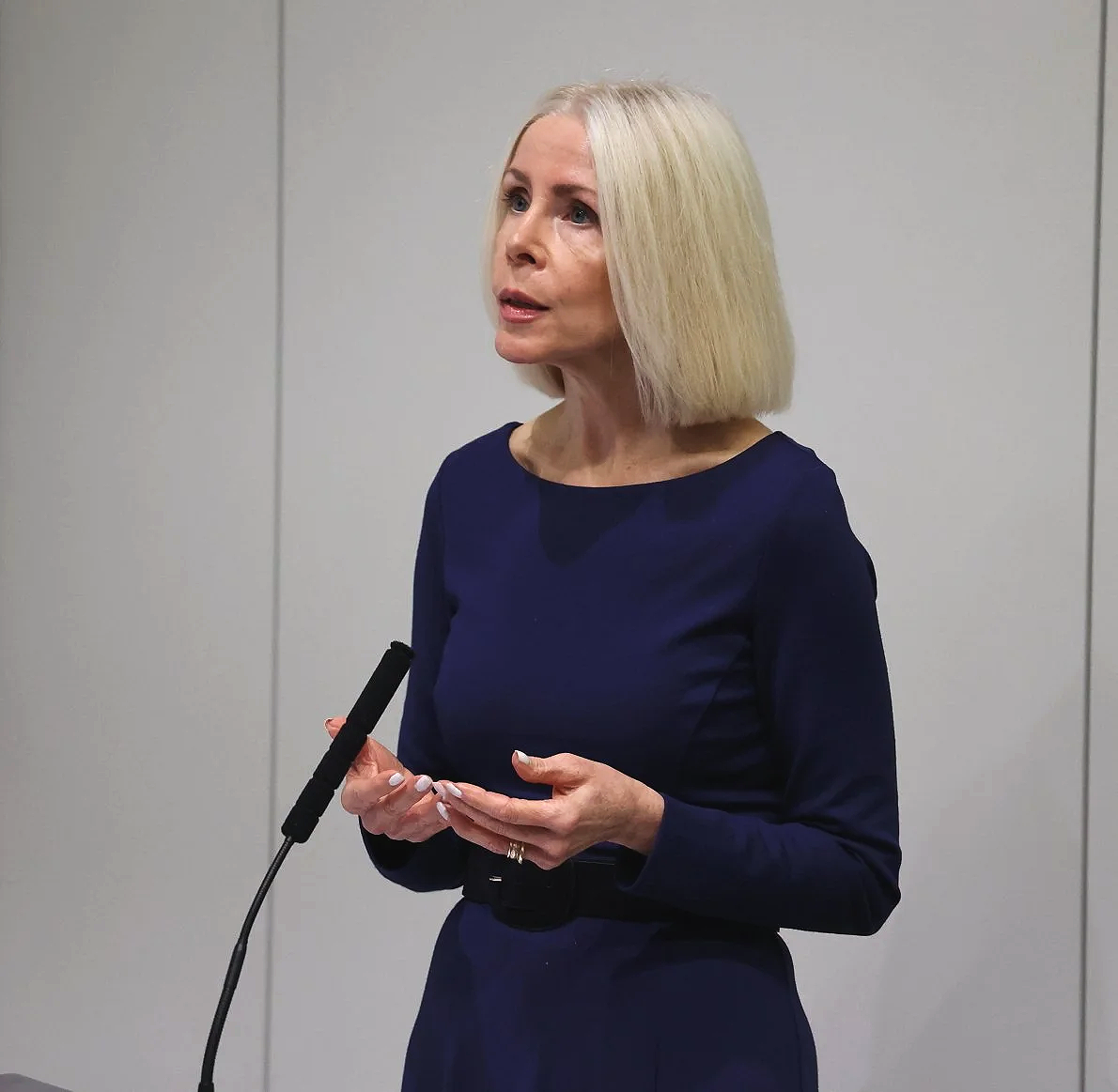 A woman with shoulder-length blonde hair, wearing a dark blue dress, stands in front of a plain gray wall, speaking into a microphone.