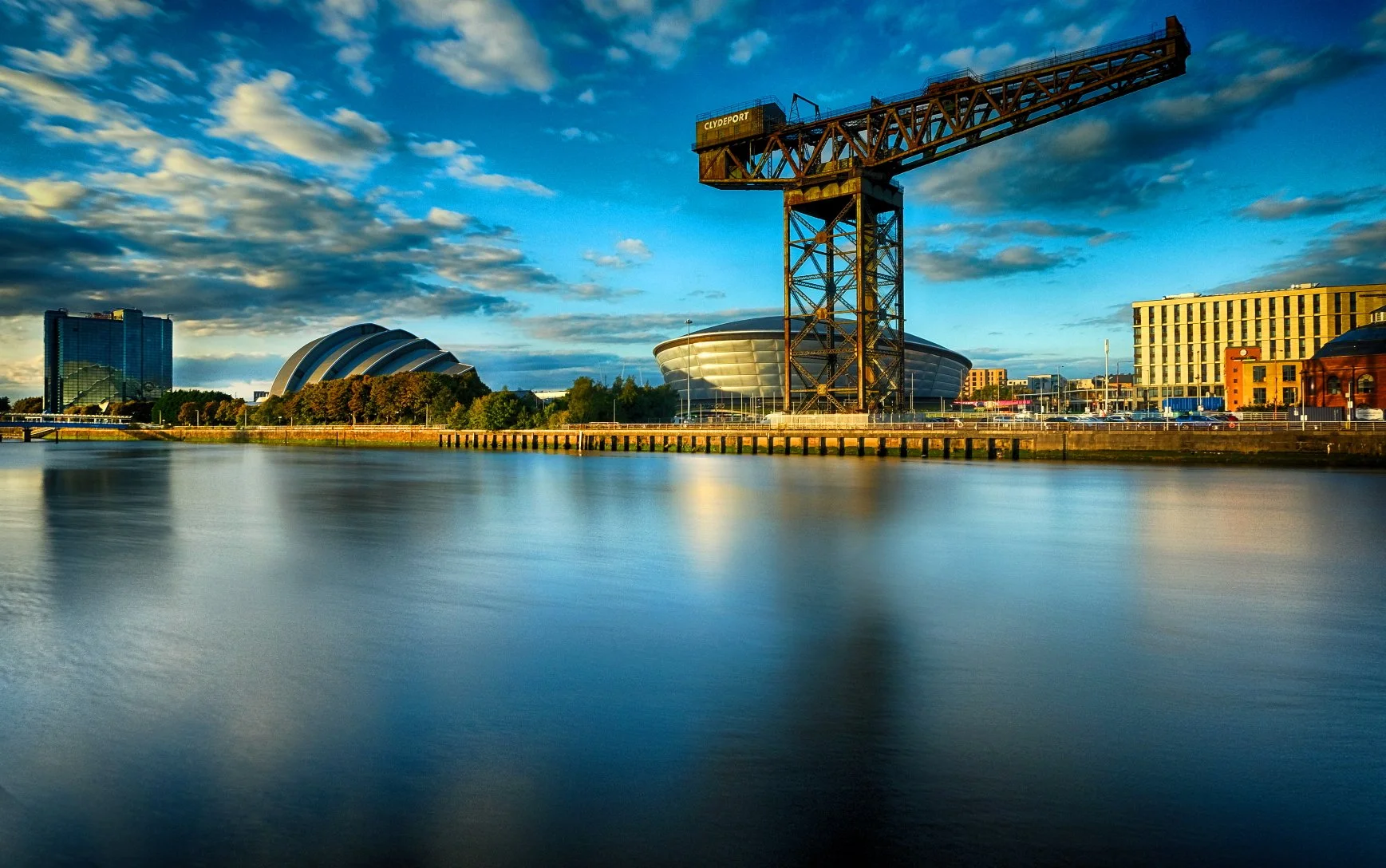 A cityscape of downtown Glasgow, Scotland, featuring the Finnieston Crane, the Armadillo concert hall, and the SSE Hydro arena, with reflections on the water in the foreground and a partly cloudy sky overhead.