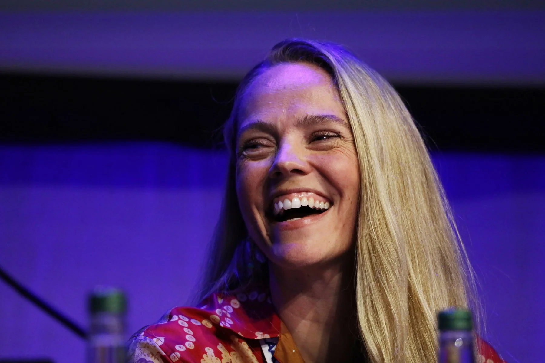 A woman with long blonde hair smiling and laughing at an indoor event, wearing a red patterned blouse with a background of purple and black.
