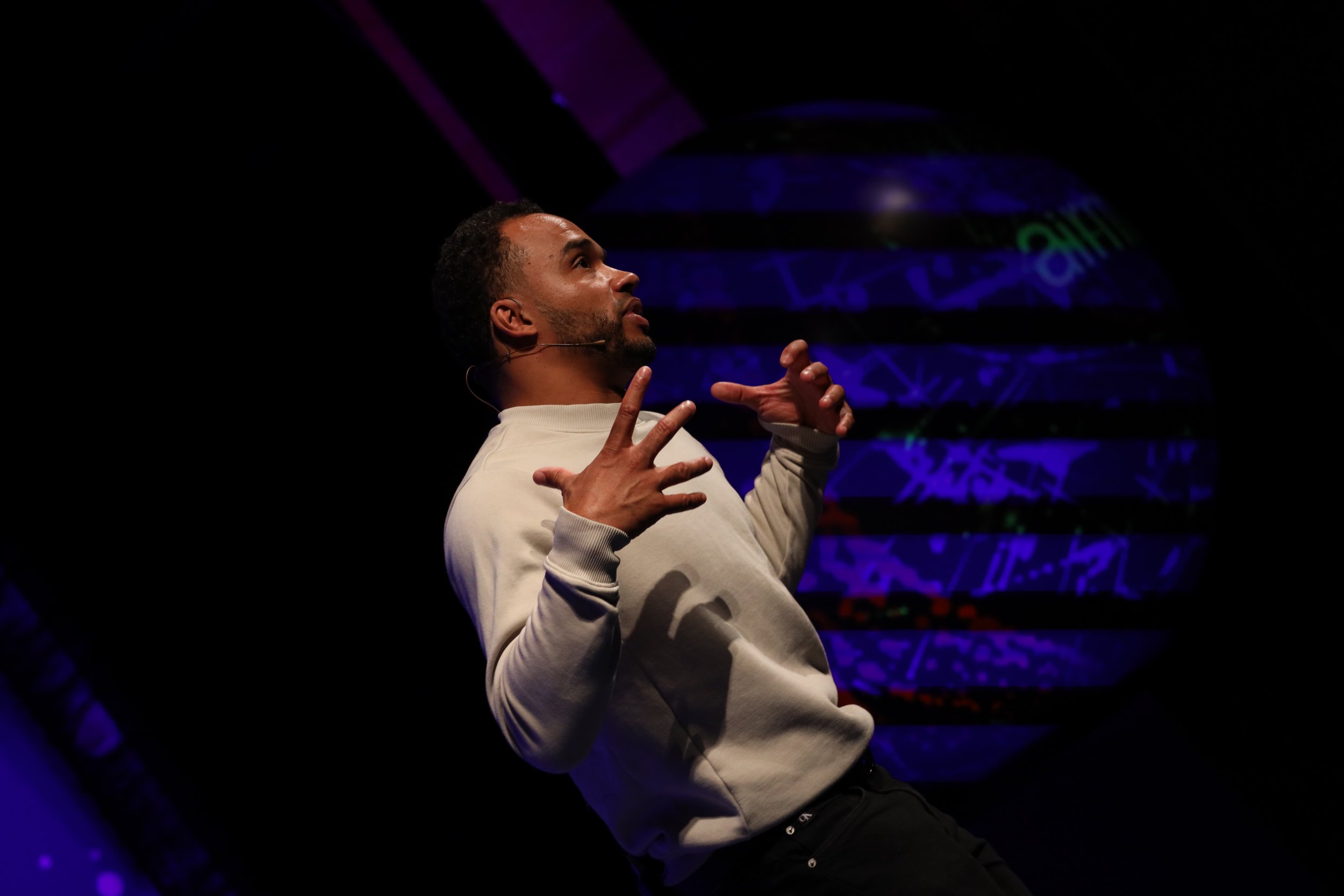 A man giving a presentation or talk on stage, gesturing with both hands, dark background with a purple and blue spherical graphic