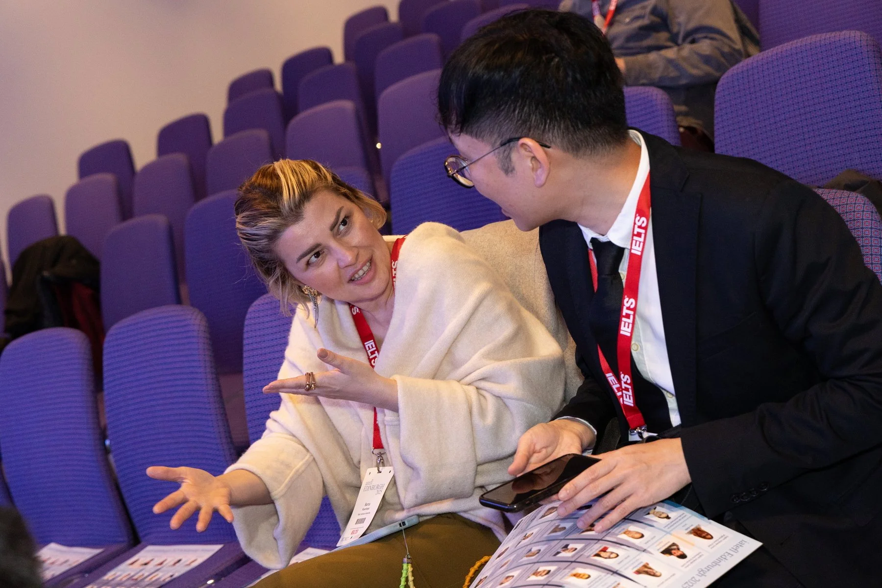 Two people are seated and engaged in conversation in an auditorium with purple seats. The woman, wearing a beige shawl and face mask hanging below her chin, is gesturing with her hands. The man, in a black suit and tie, is holding a phone and looking at her. They are both wearing conference badges with red lanyards.