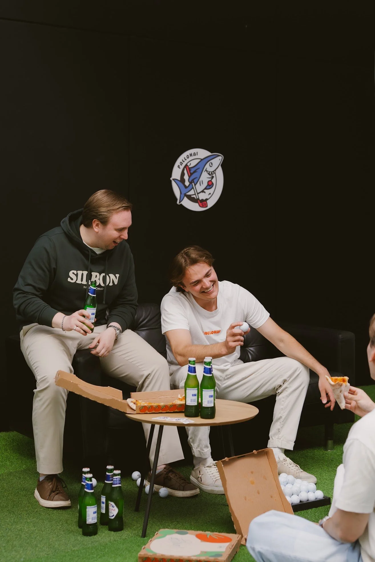 Kaksi nuorta miestä istuvat sohvalla, nauravat ja juttelevat. Toisella miehellä on olutpullo, hänellä on myös pizza, ja pizzeria ja tennisaiheinen logo seinällä taustalla.