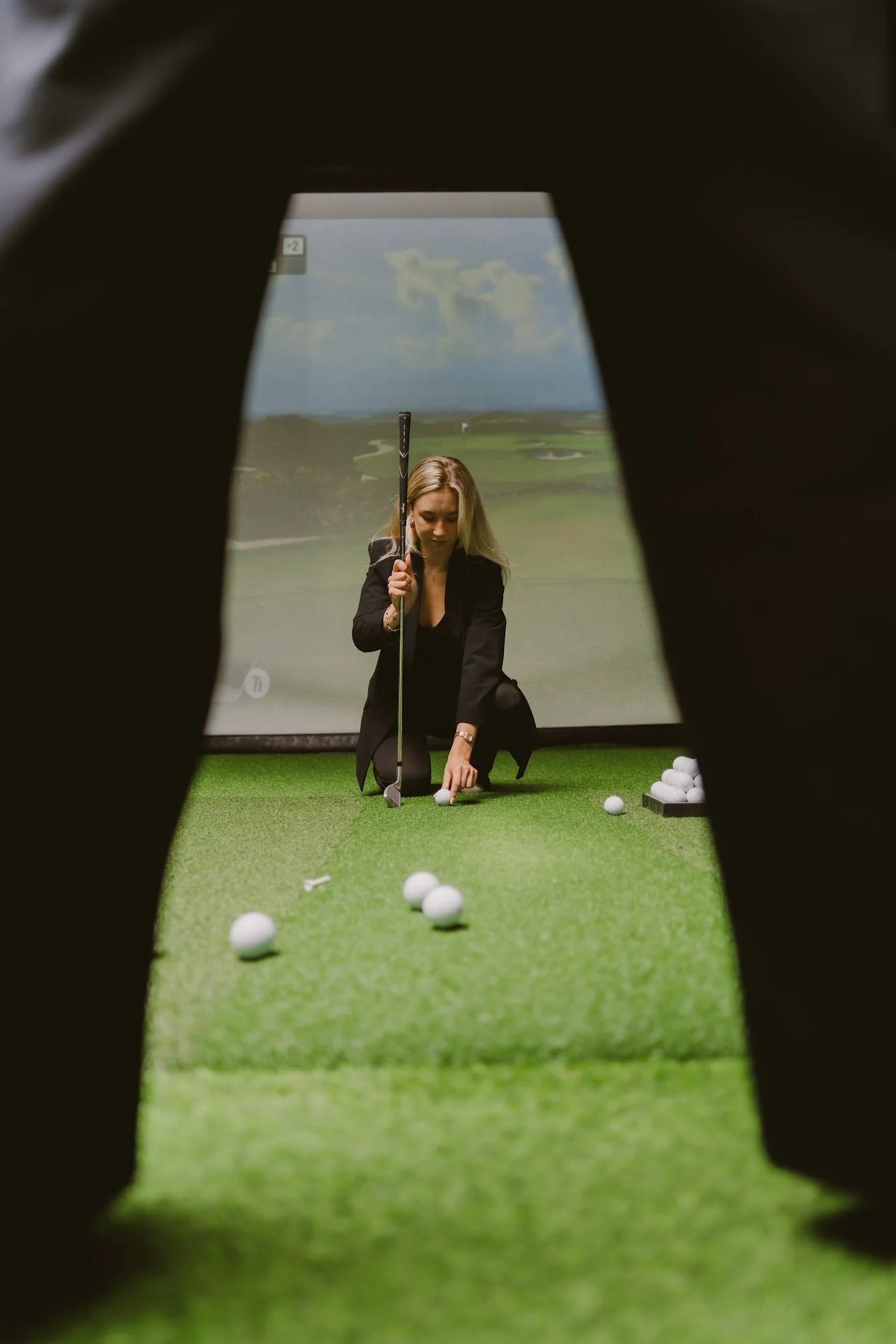 Nainen treenaa golf-klubilla, katse alaspäin palloon, taustalla golfkenttä ja pilvinen taivas