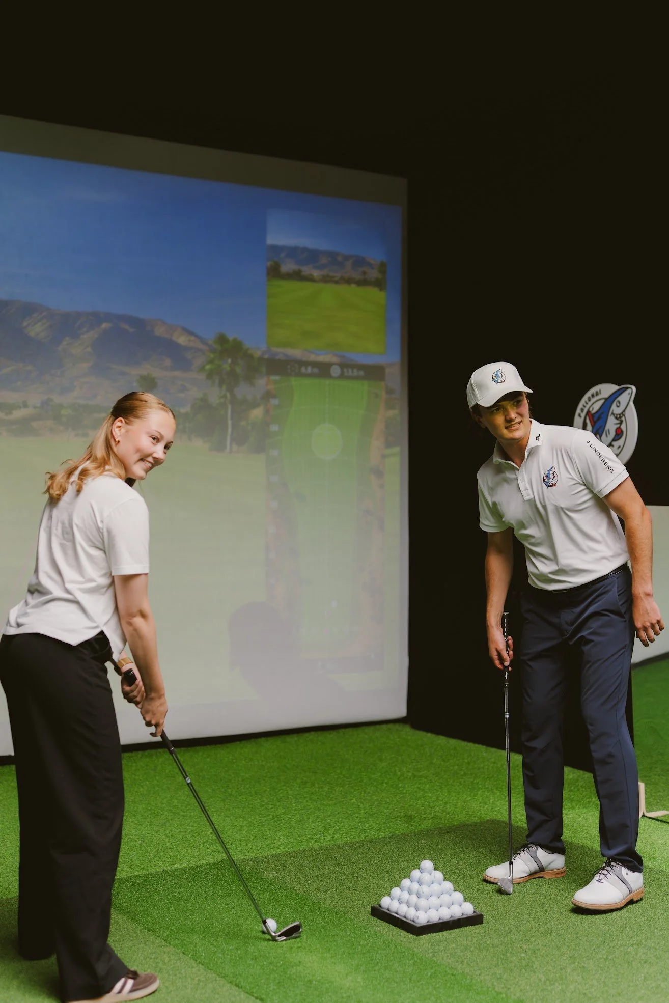 Kaksi ihmistä harjoittelee golfia sisäkoulussa. Taustalla suuri näyttö ja golfmaali.