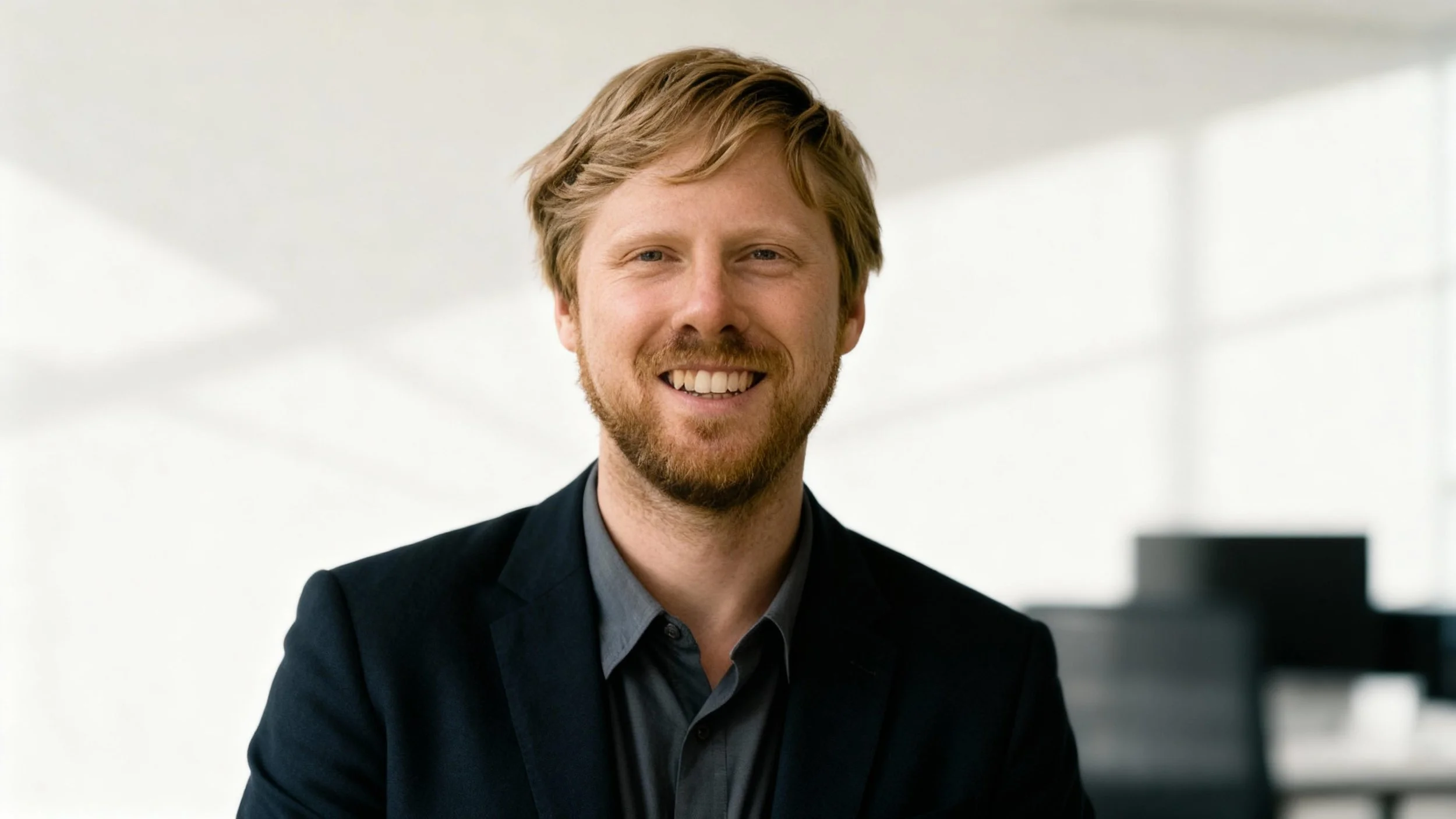A smiling man with light brown hair and a beard, wearing a dark blazer and a gray shirt, standing in a modern office with a bright background.