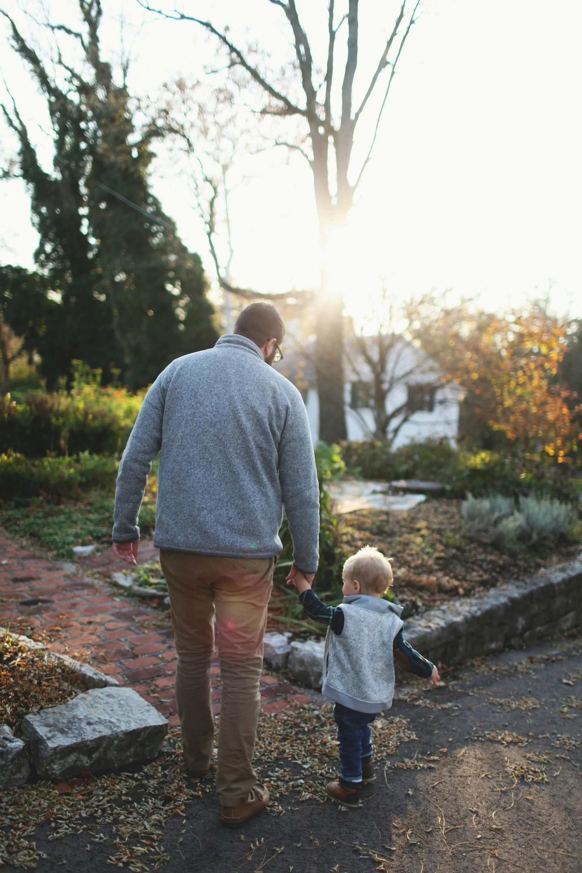 A man and a young child walking hand in hand on a garden path during sunset.