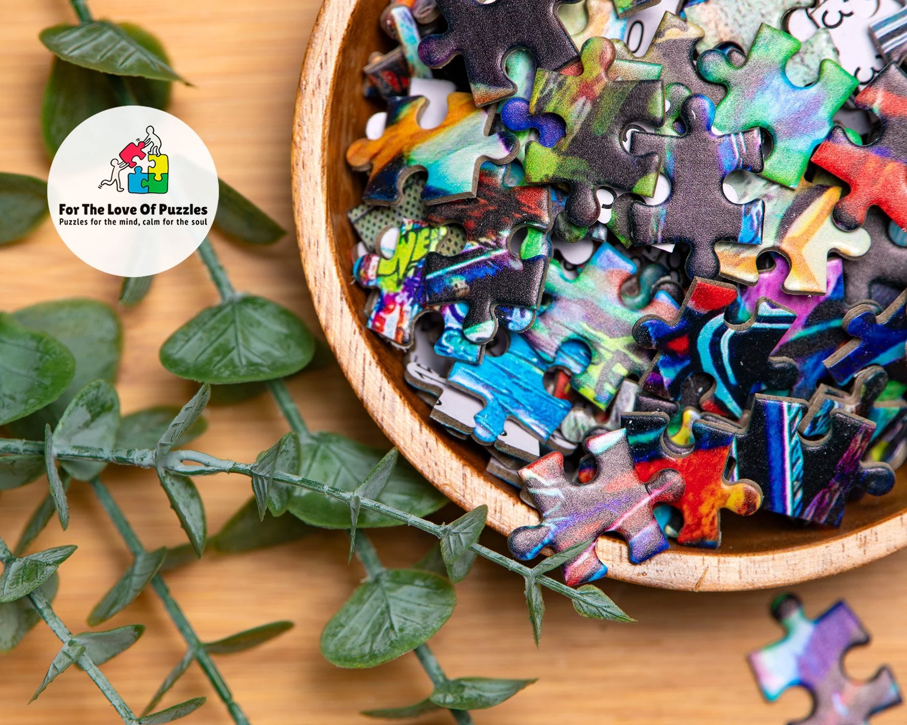 Colorful jigsaw puzzle pieces in a wooden bowl on a wooden surface with green leafy plant nearby.