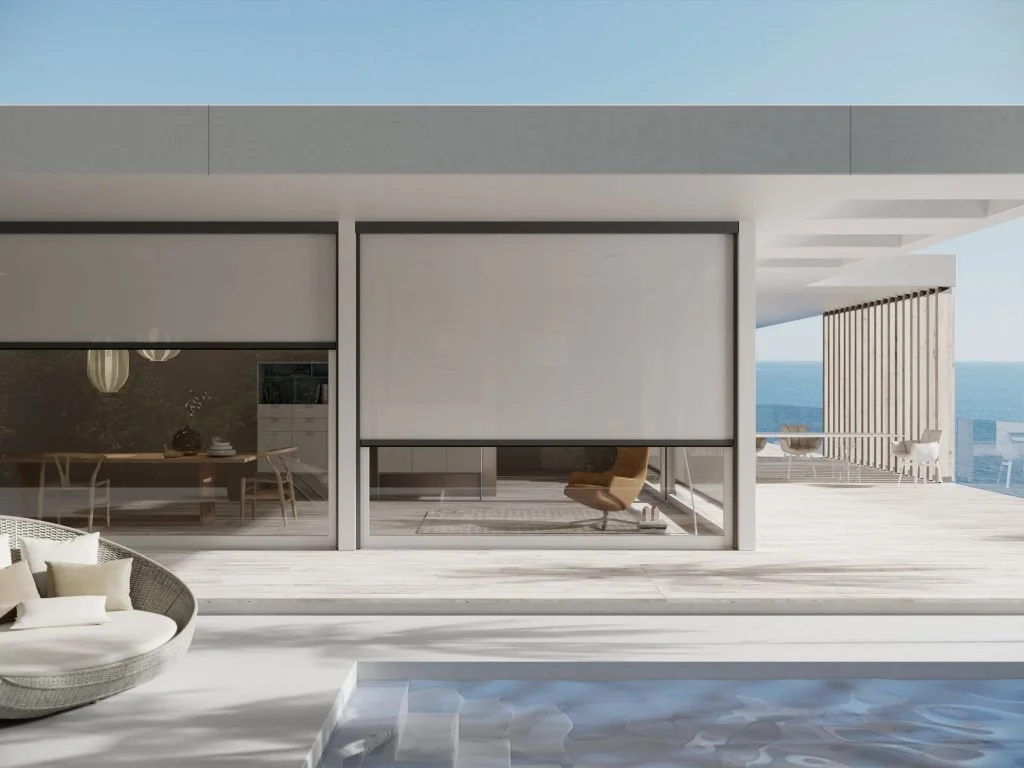 Modern house with large glass windows overlooking the ocean. Outdoor patio with seating area and wooden slat privacy screen, with a pool in the foreground.