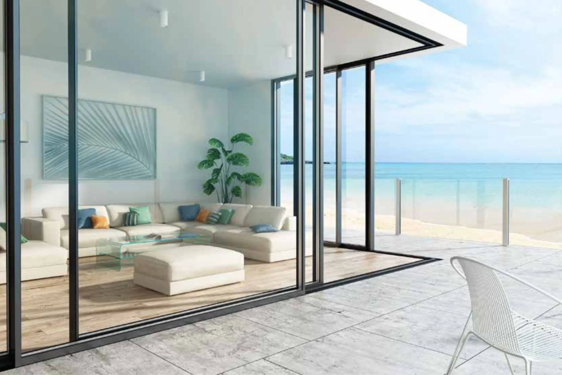 Living room with glass walls overlooking a beach, featuring a white L-shaped sofa with colorful pillows, a glass coffee table, a large green plant, and beach scenery outside.
