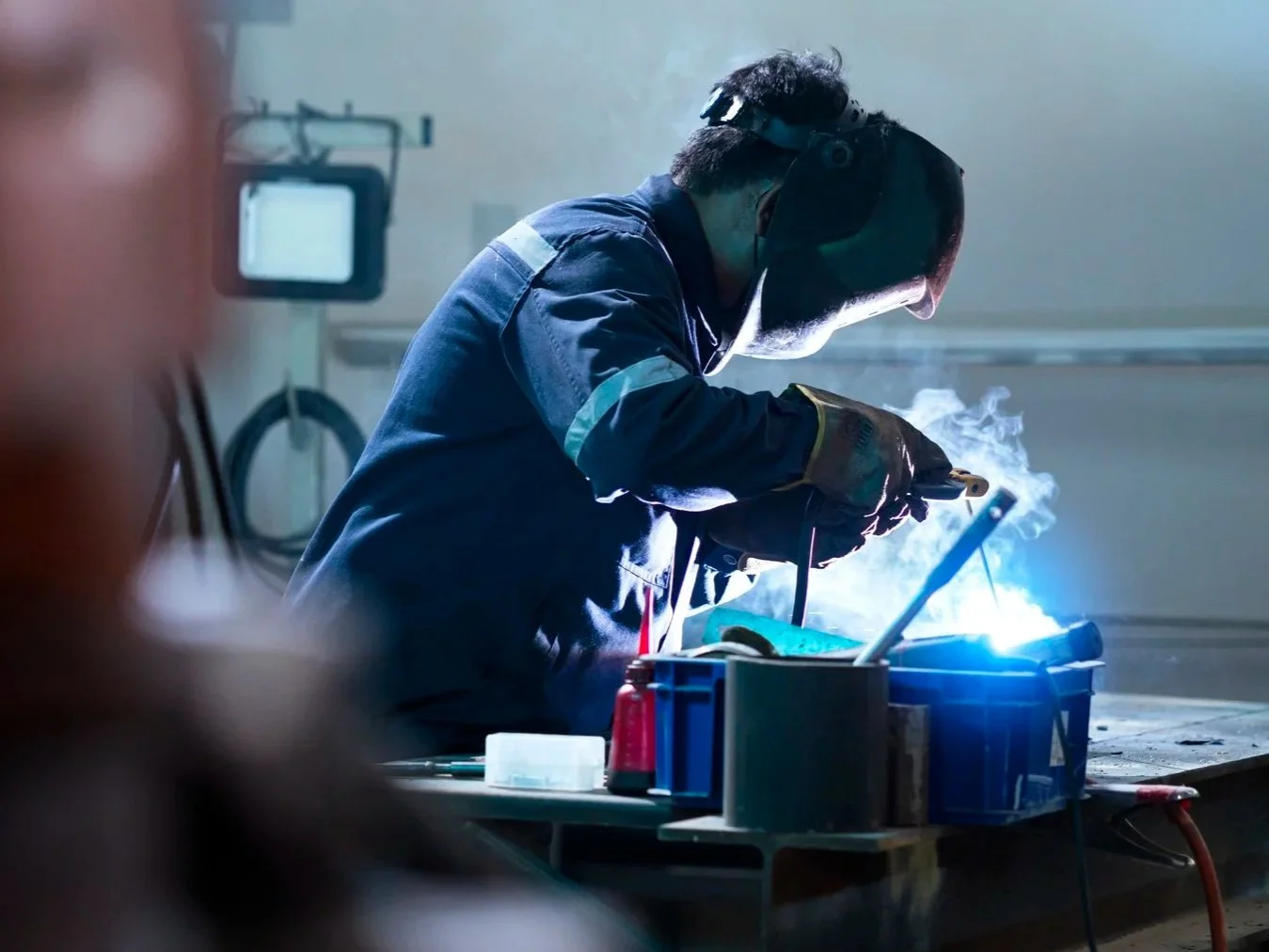A person in protective gear welding metal in a workshop, with sparks and smoke visible.