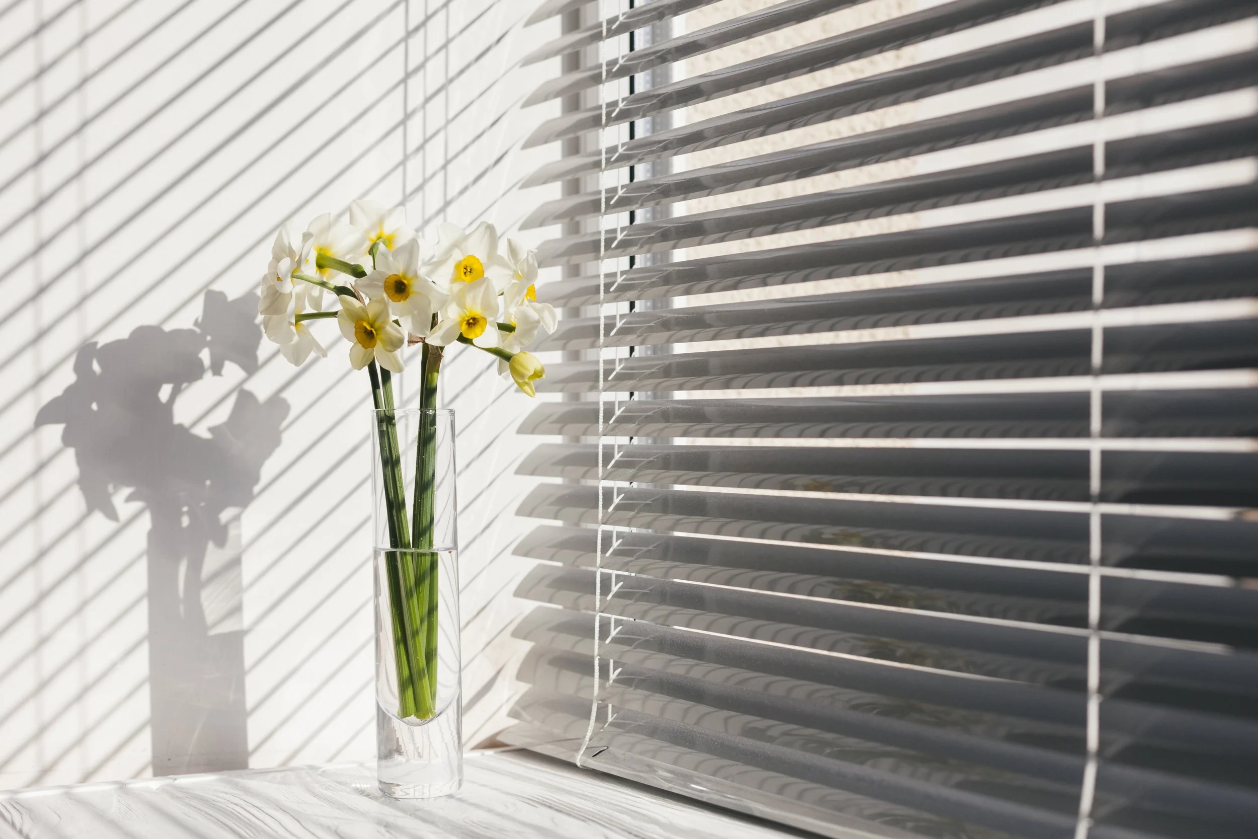 A clear glass vase with white and yellow flowers on a white windowsill, with sunlight casting shadows of the flowers and window blinds on the wall.
