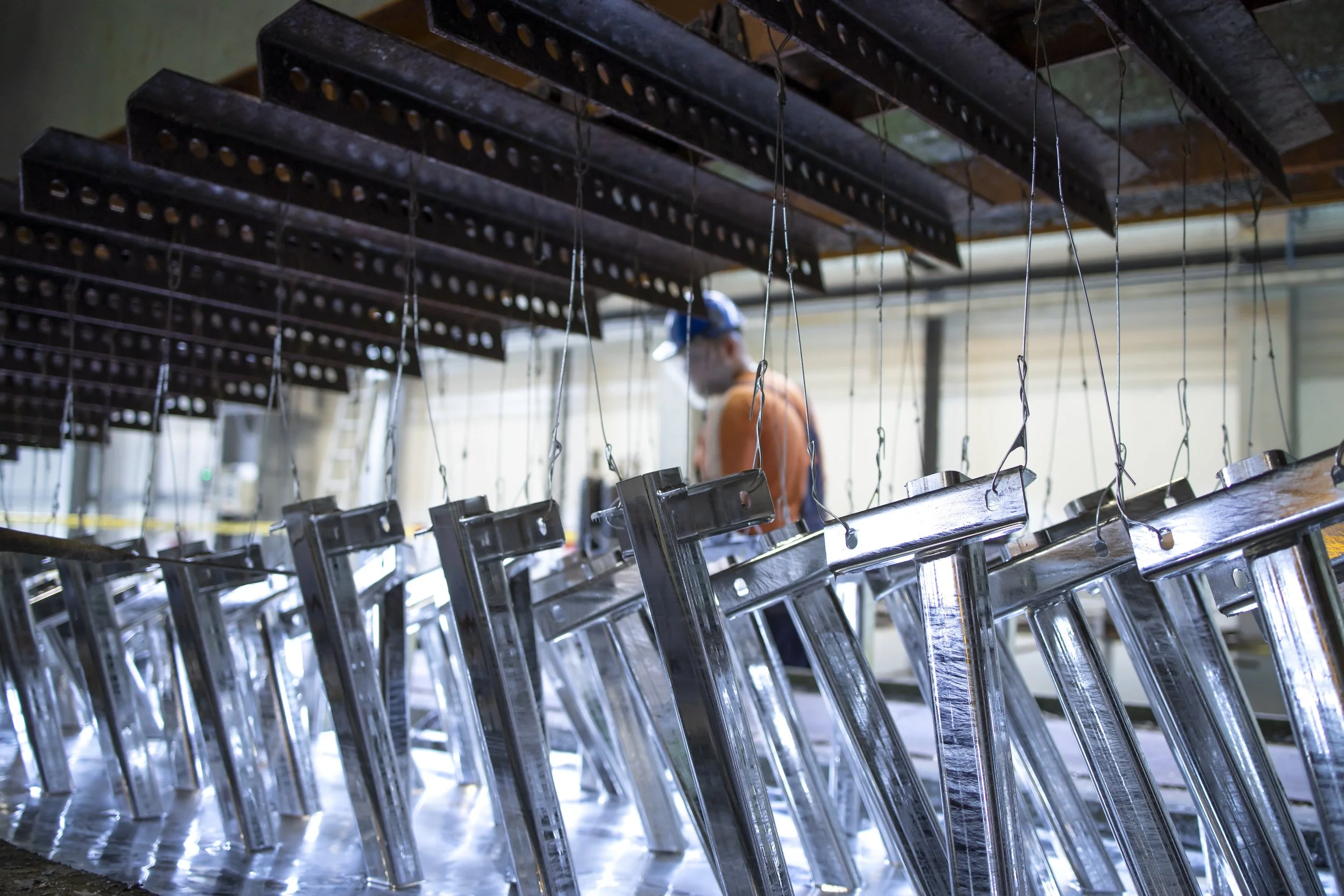 Metal chair legs hanging upside down in a manufacturing workshop with a worker in the background wearing a mask, cap, and safety vest.
