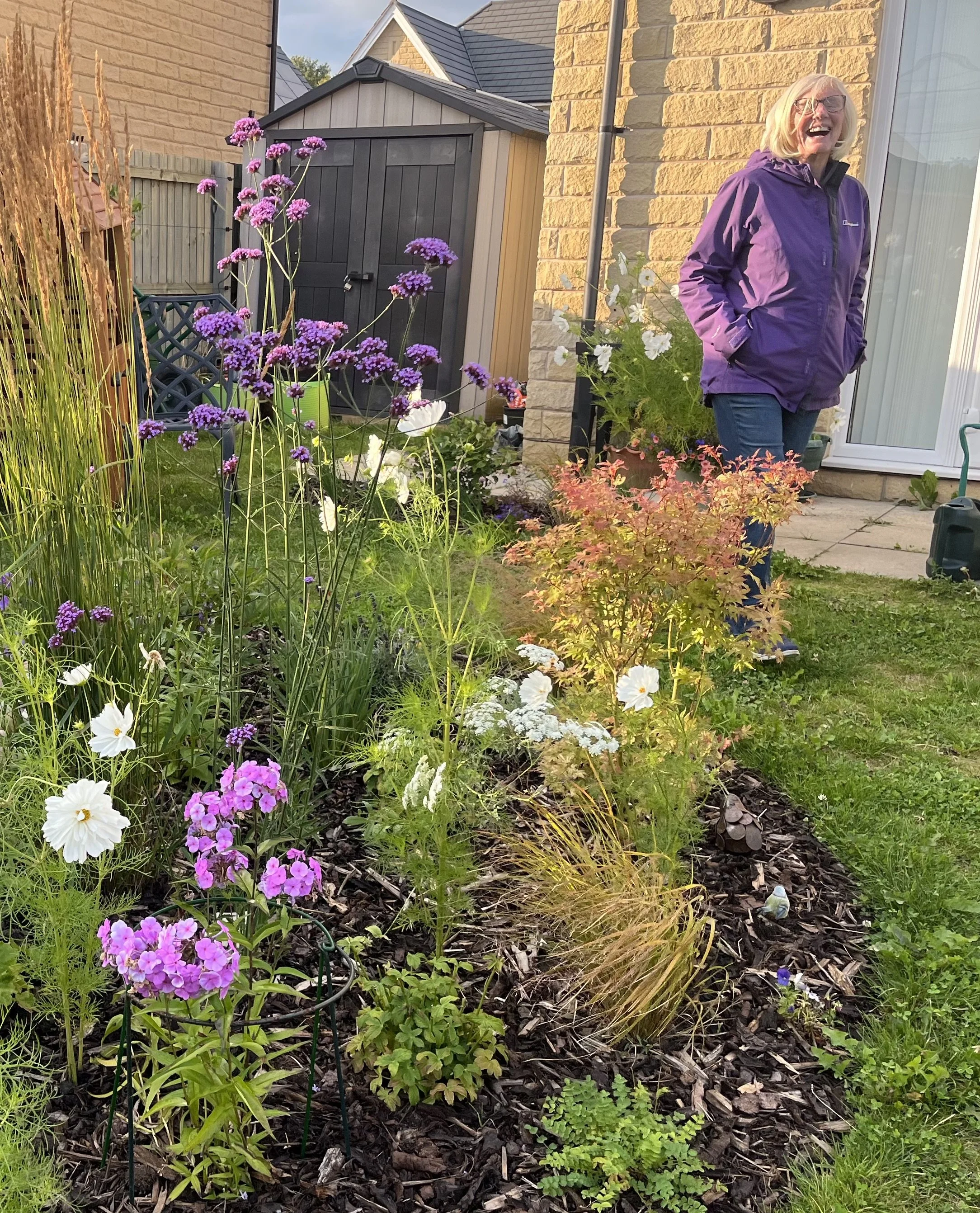 A woman with blonde hair and glasses wearing a purple jacket stands outside smiling in a garden with colorful flowers, a shed, and a house in the background.