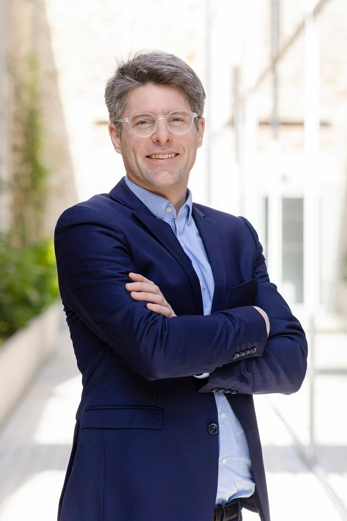 Un homme d'affaires souriant portant des lunettes, vêtu d'une veste bleue et d'une chemise, debout avec les bras croisés dans un espace lumineux avec des fenêtres