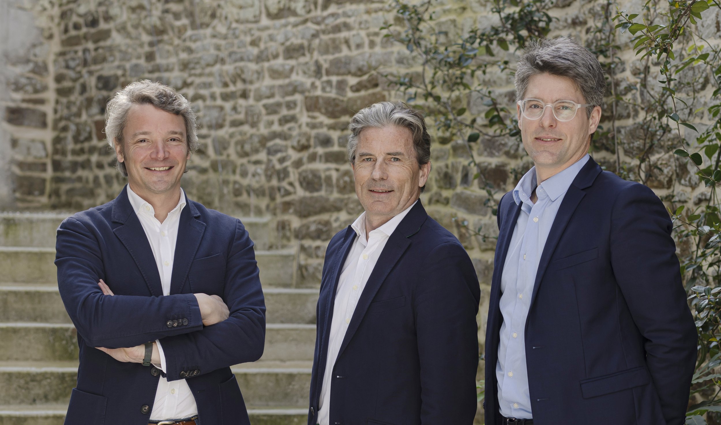 Trois hommes portant des costumes posant devant un mur en pierre et des escaliers en pierre.