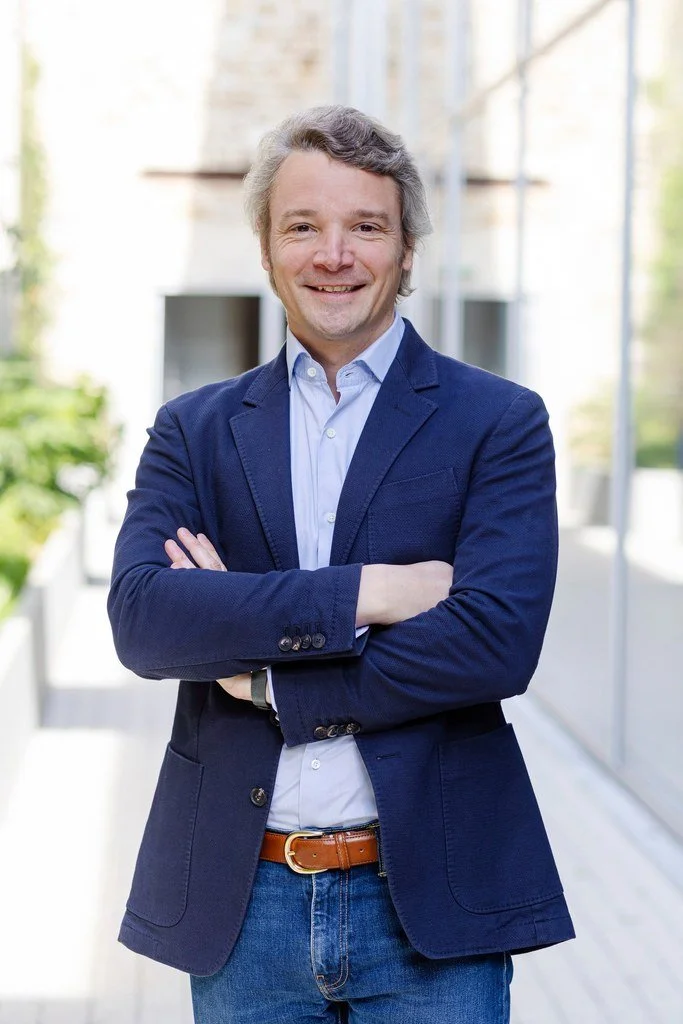 Un homme souriant, en veste bleue et chemise blanche, avec les bras croisés, dans un environnement moderne et lumineux.