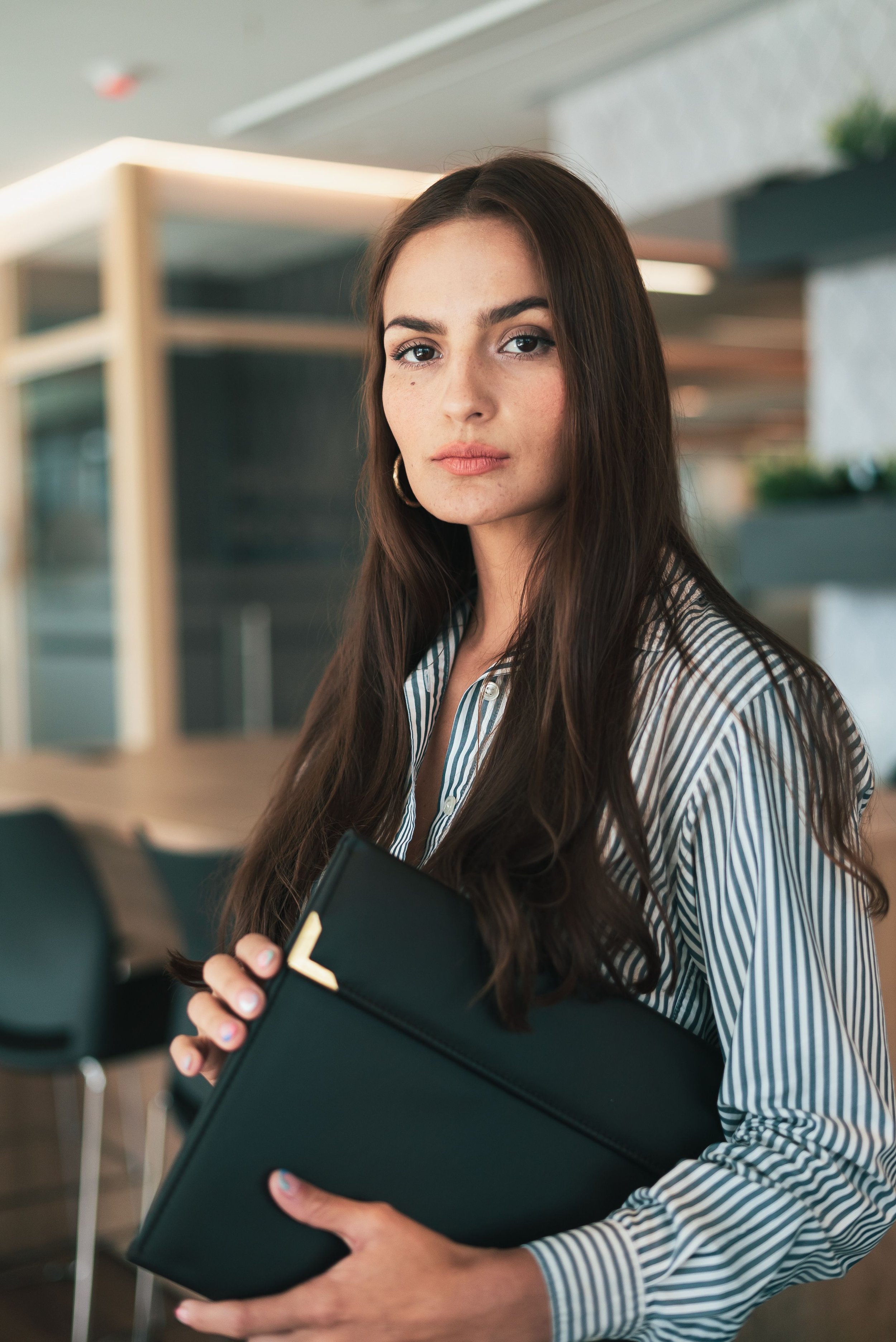 Une jeune femme aux cheveux longs et bruns portant une chemise à rayures blanches et bleues, tenant un cahier noir avec des coins dorés dans un environnement intérieur moderne.