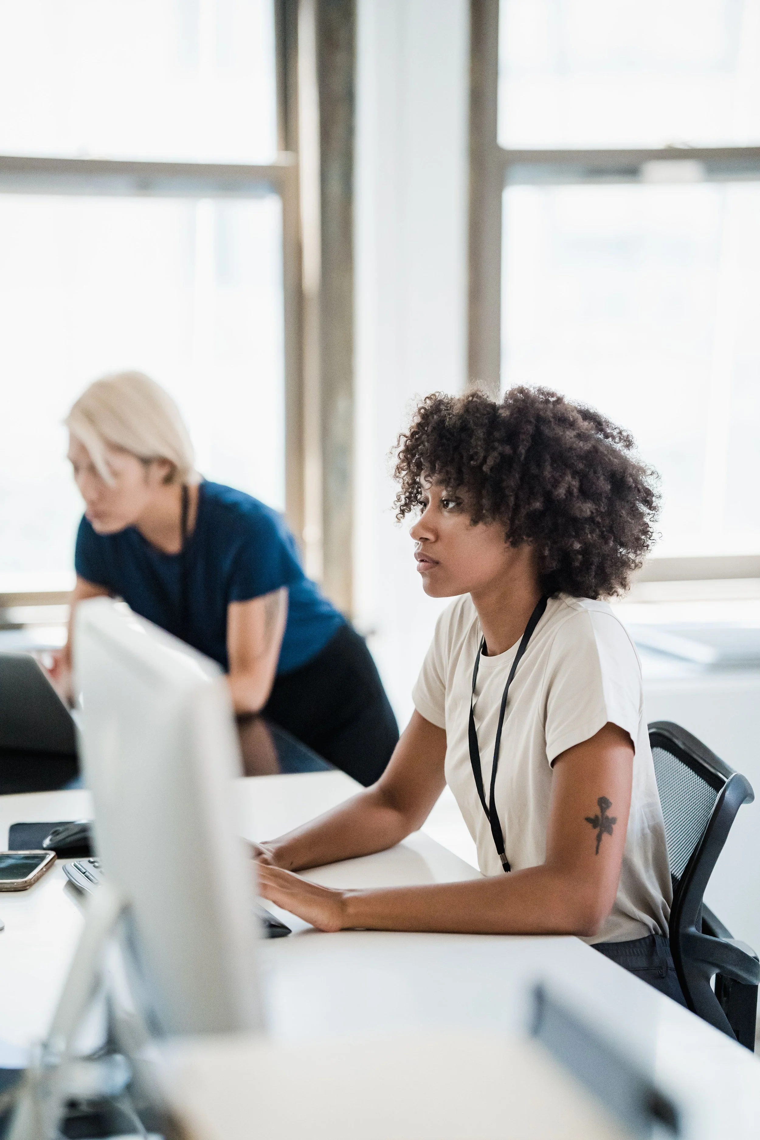 Deux femmes assises à une table dans un bureau, travaillant sur des ordinateurs, devant une grande fenêtre with un éclairage naturel.