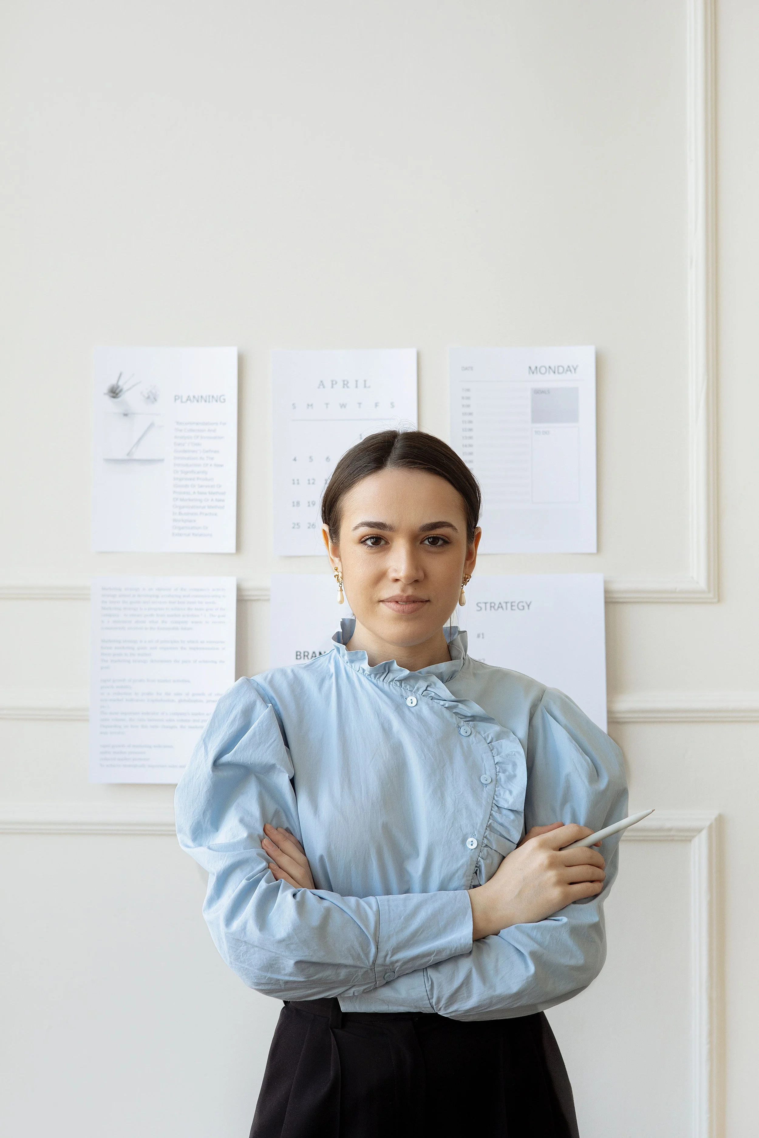 Une femme souriante avec les bras croisés, portant une chemise bleu clair et des boucles d'oreilles dorées, se tient devant un tableau blanc avec des papiers affichés.