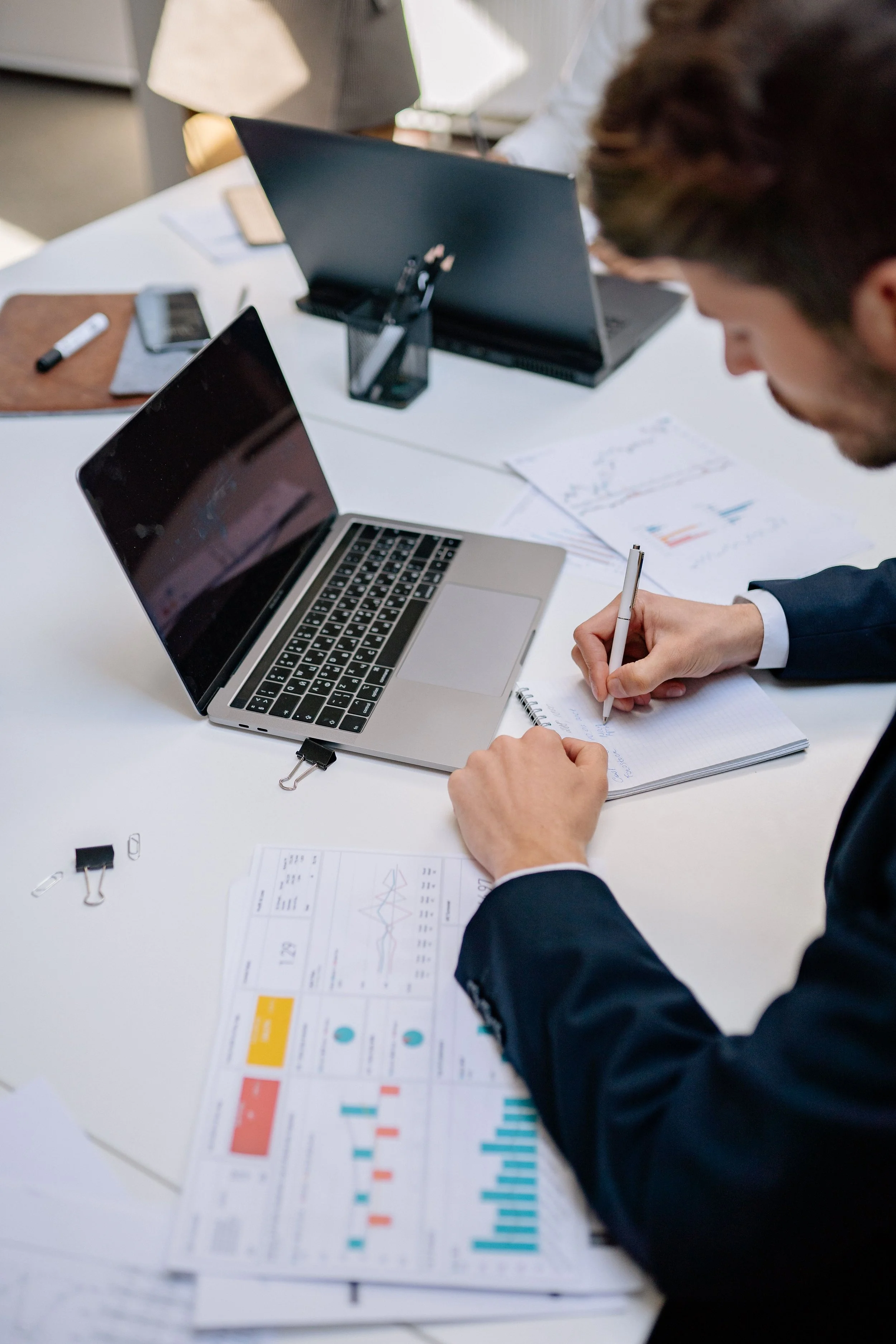 Une personne en costume travail, écrivant sur un cahier avec des graphiques, à côté d'un ordinateur portable et des documents avec des graphiques colorés, dans un bureau lumineux.