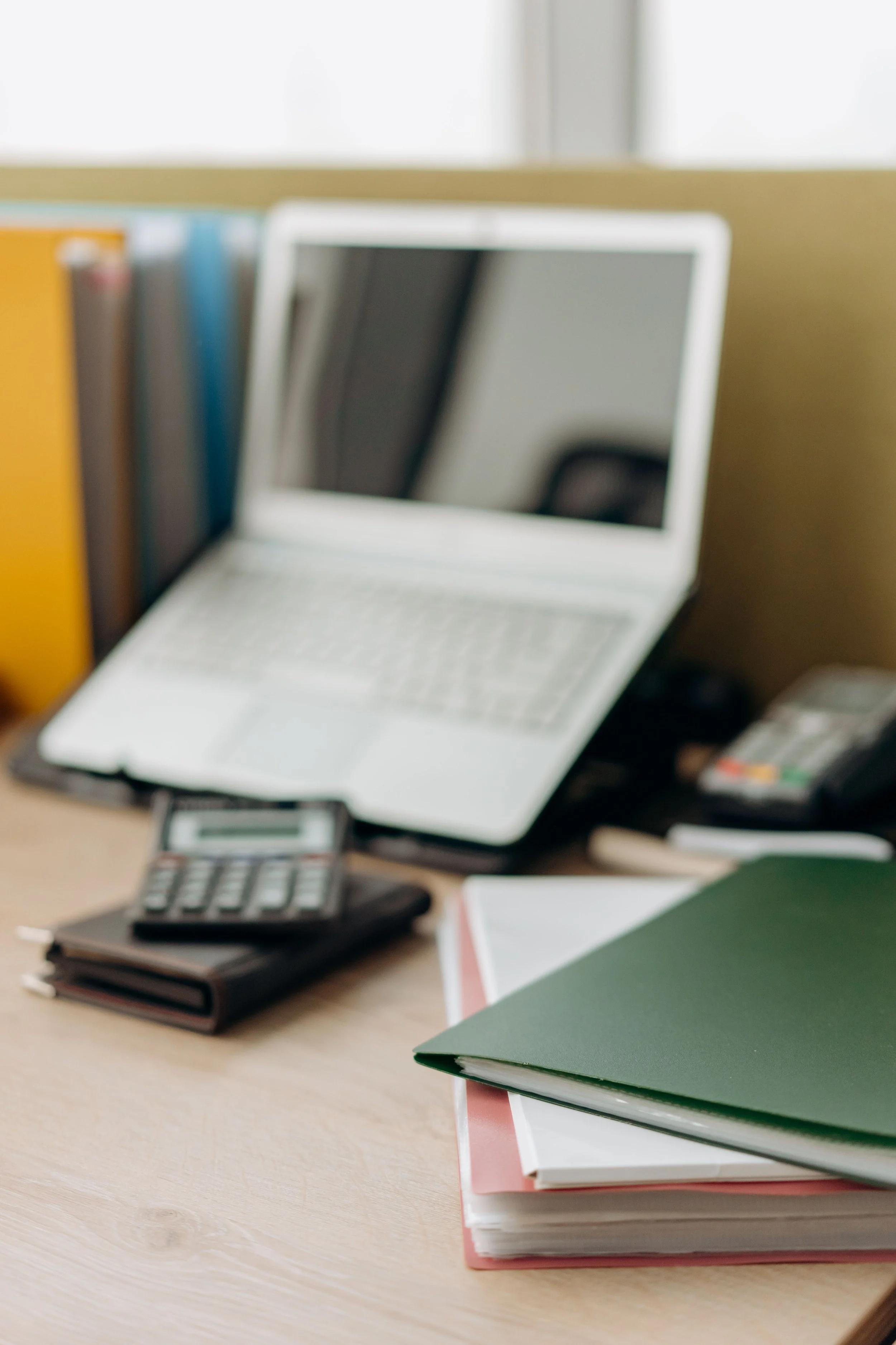 Un bureau avec un ordinateur portable, plusieurs classeurs empilés, un calculatrice, et des cahiers ou carnets.