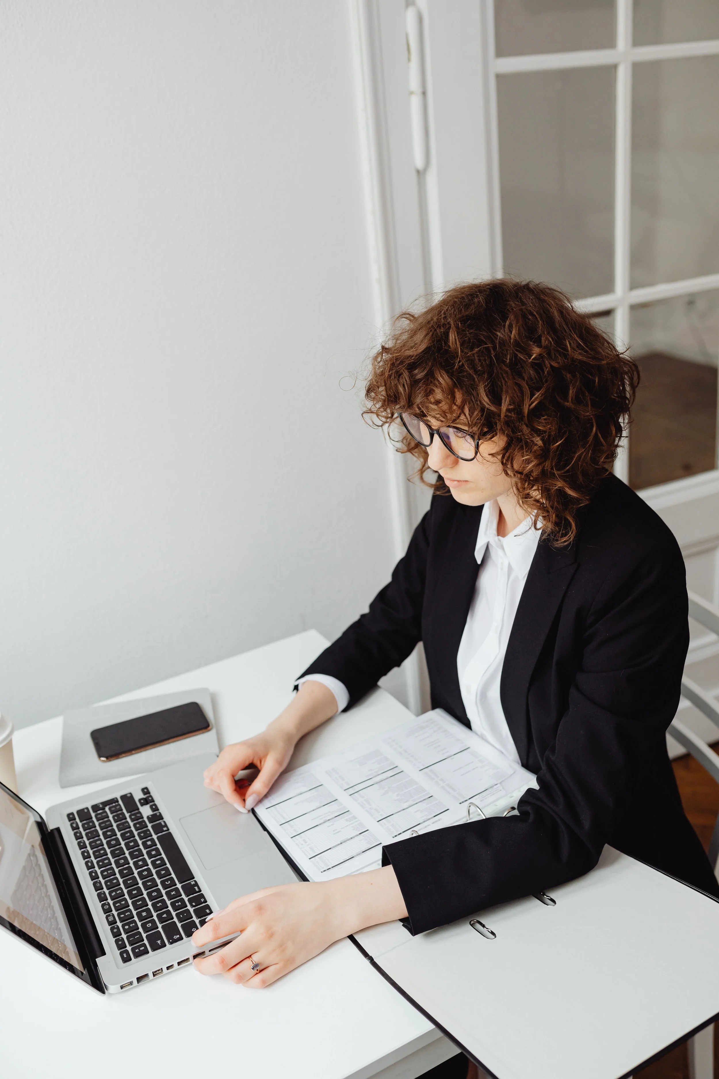 Une femme portant un blazer noir et des lunettes travaille à un bureau blanc, utilisant un ordinateur portable, avec un téléphone et des documents organisés devant elle. Elle semble concentrée sur sa tâche.