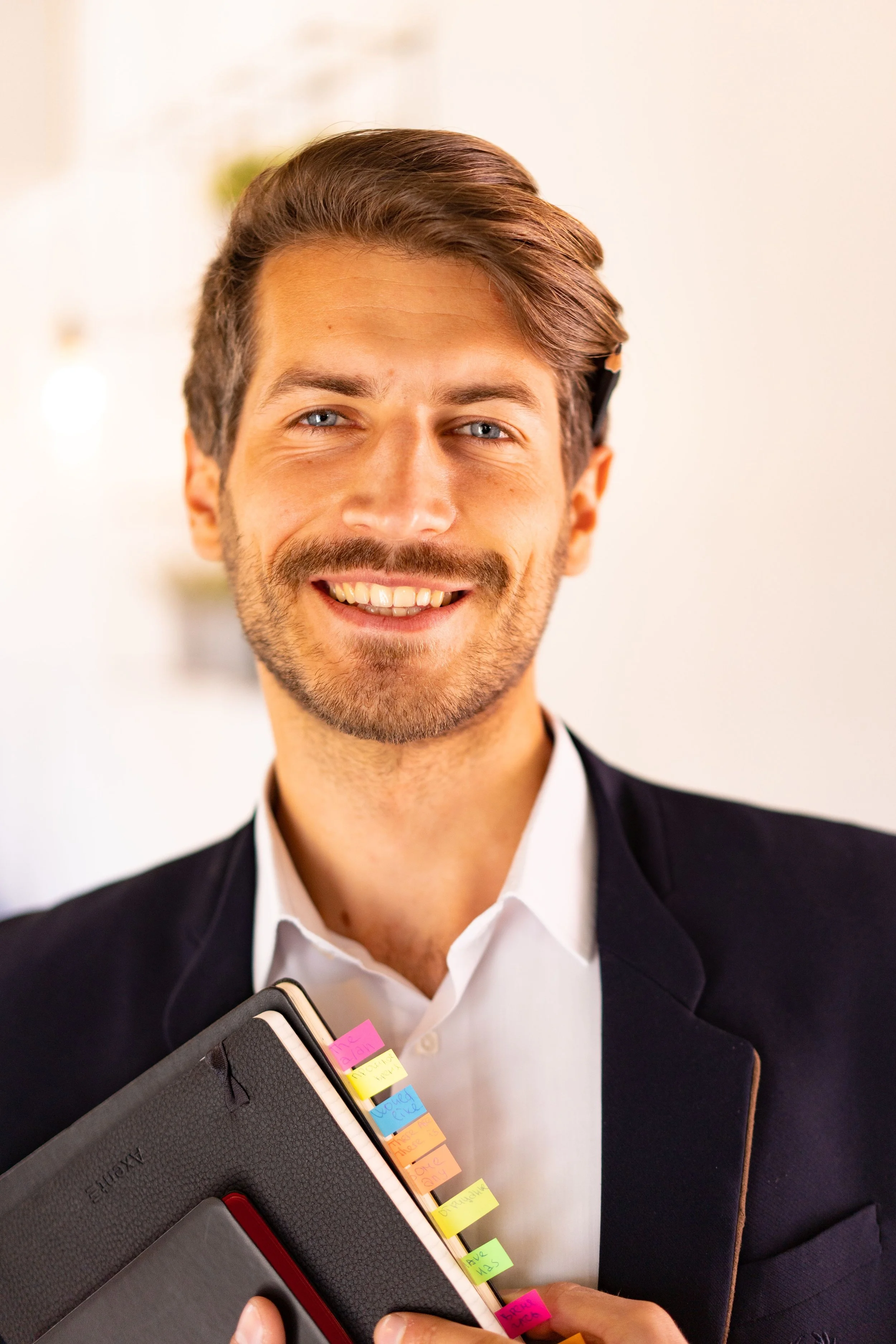 Un homme souriant avec des cheveux bruns et une barbe porte un costume noir avec une chemise blanche, tenant un carnet noir avec des post-it colorés attachés.