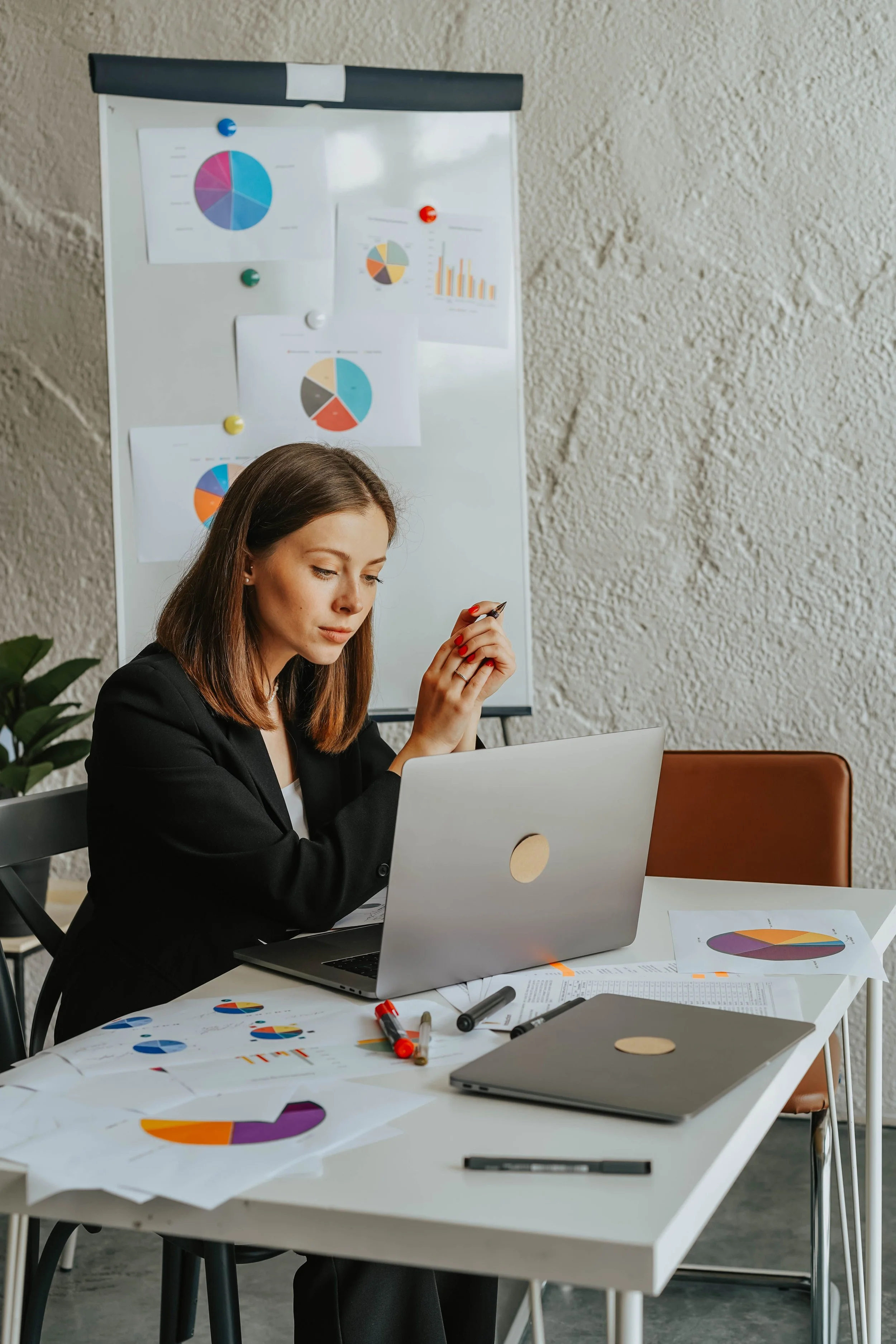 Femme d'affaires travaillant à un bureau avec un ordinateur portable, des papiers avec des graphiques, des marqueurs, et un tableau blanc avec des graphiques colorés au fond.